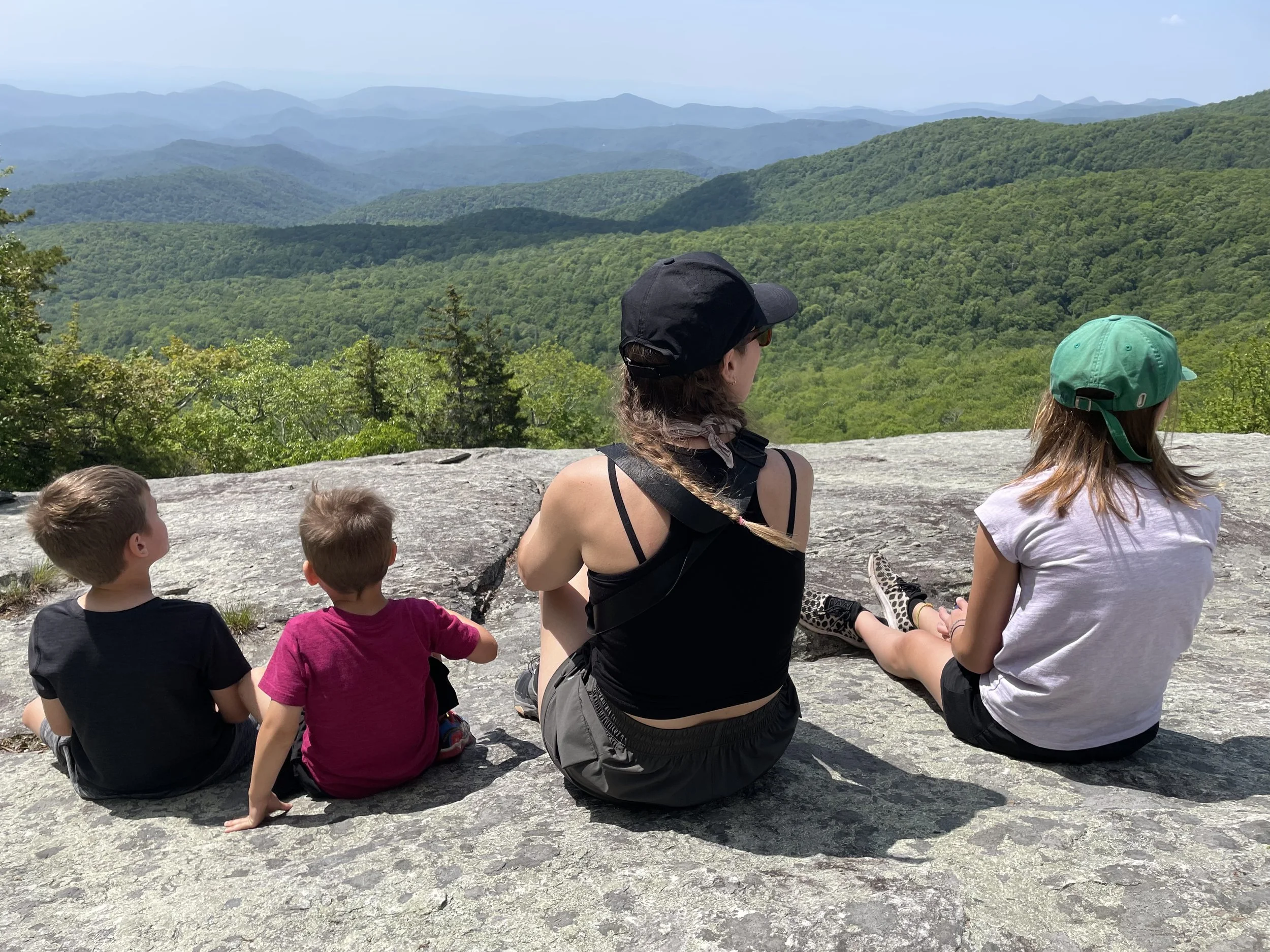 A group of children and a woman sitting on a large rock, overlooking a mountain range with lush green forests and rolling hills.