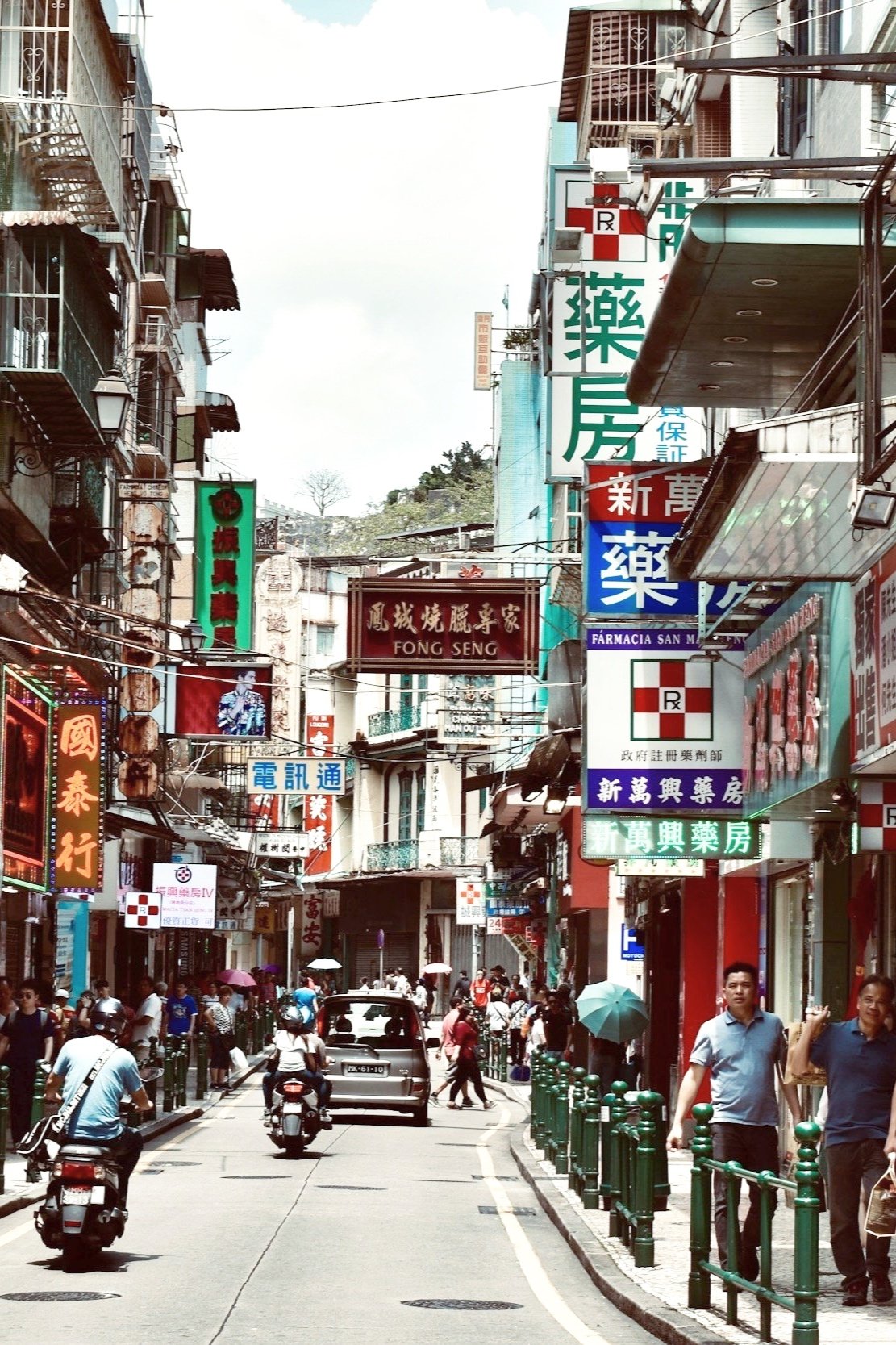 A busy city street in an Asian city with numerous colorful signs in Chinese and English, people walking, some with umbrellas, cars and scooters on the road, buildings with balconies and various storefronts.