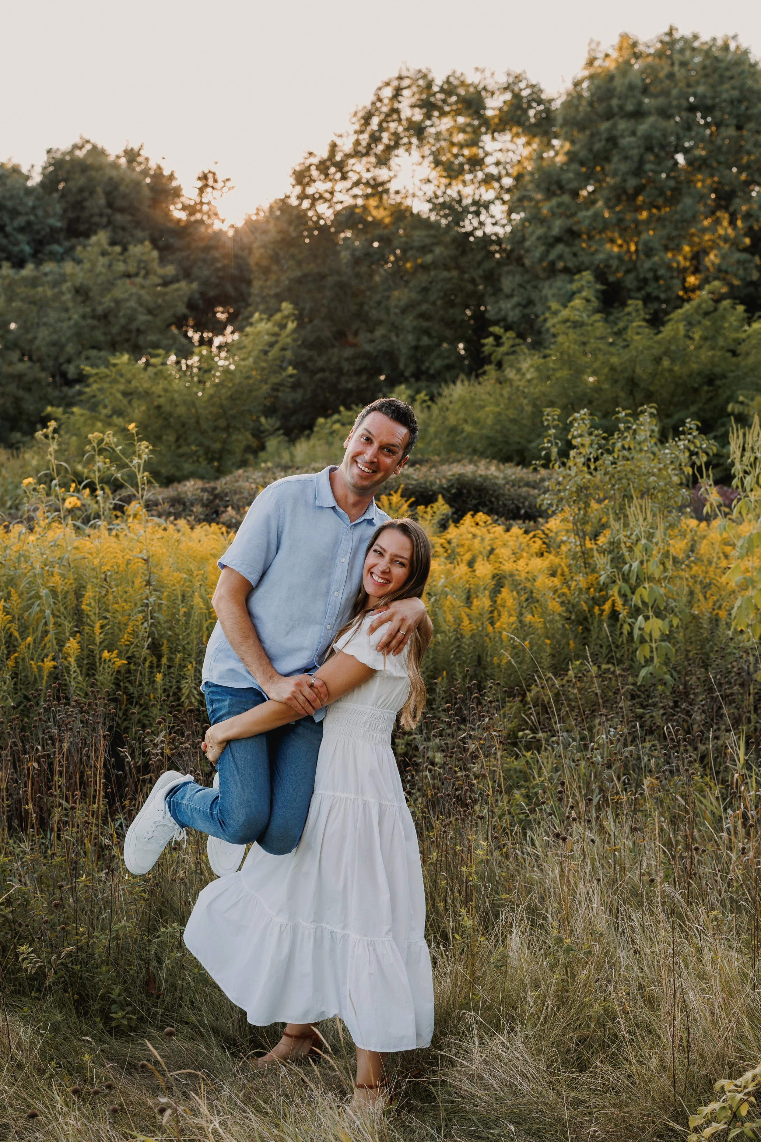 A joyful man and woman in a field of yellow flowers, with trees and the setting sun in the background.