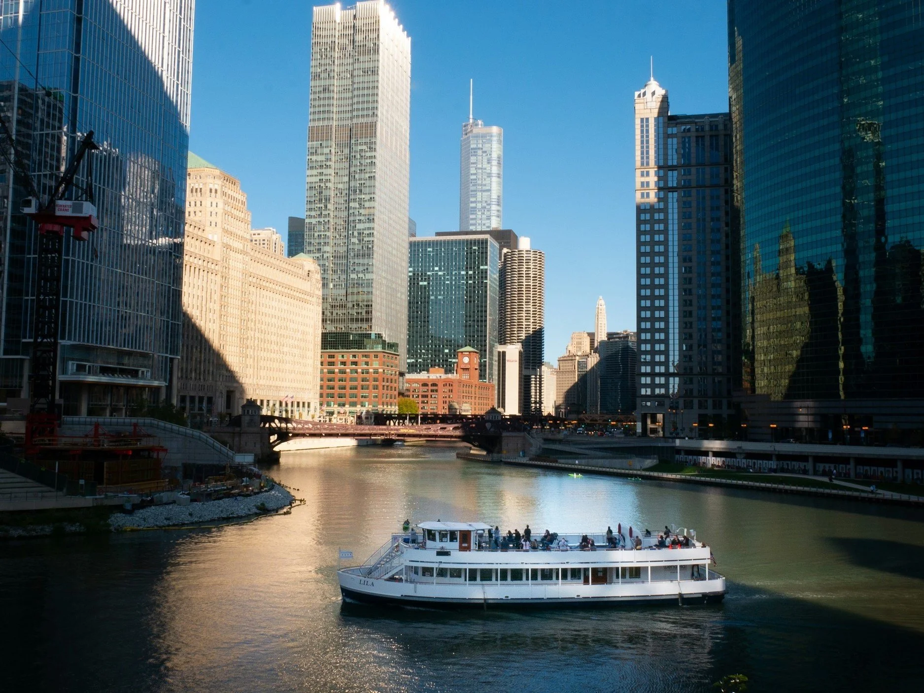 A cityscape with tall buildings and a waterway in the foreground. A boat with people is sailing on the water under a clear blue sky.