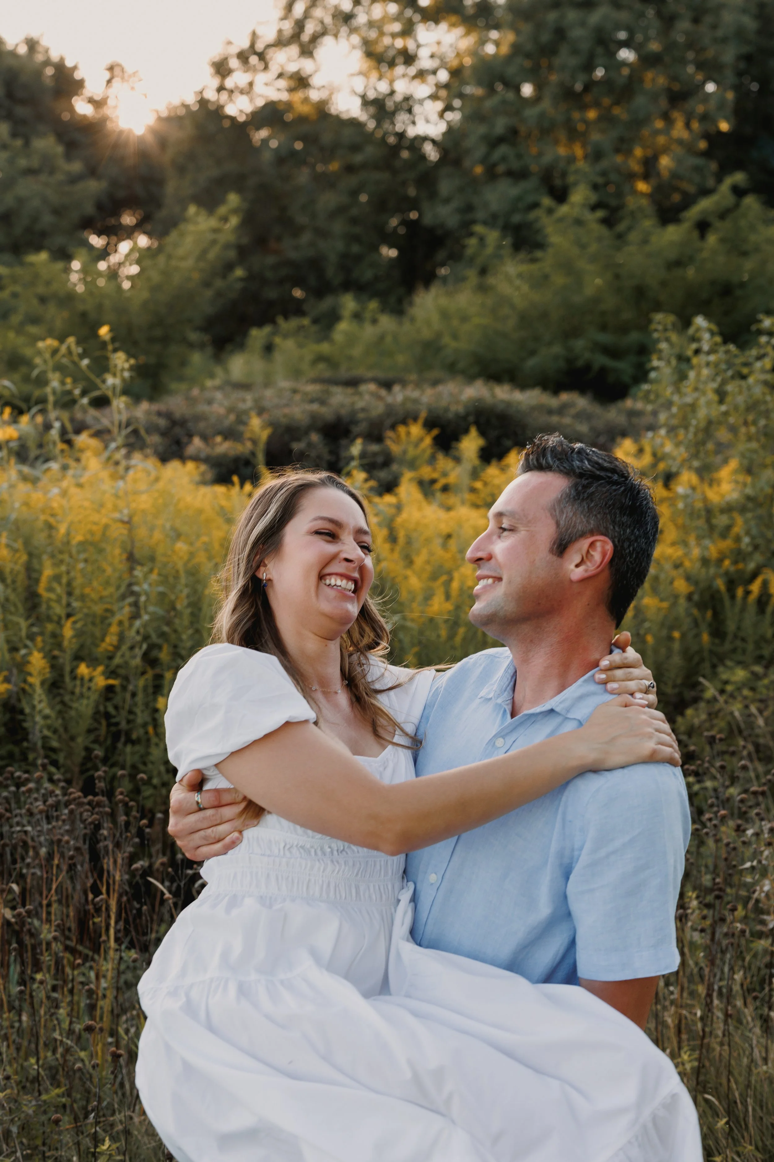 A smiling couple enjoying a hug in a field of yellow flowers with trees and the setting sun in the background.