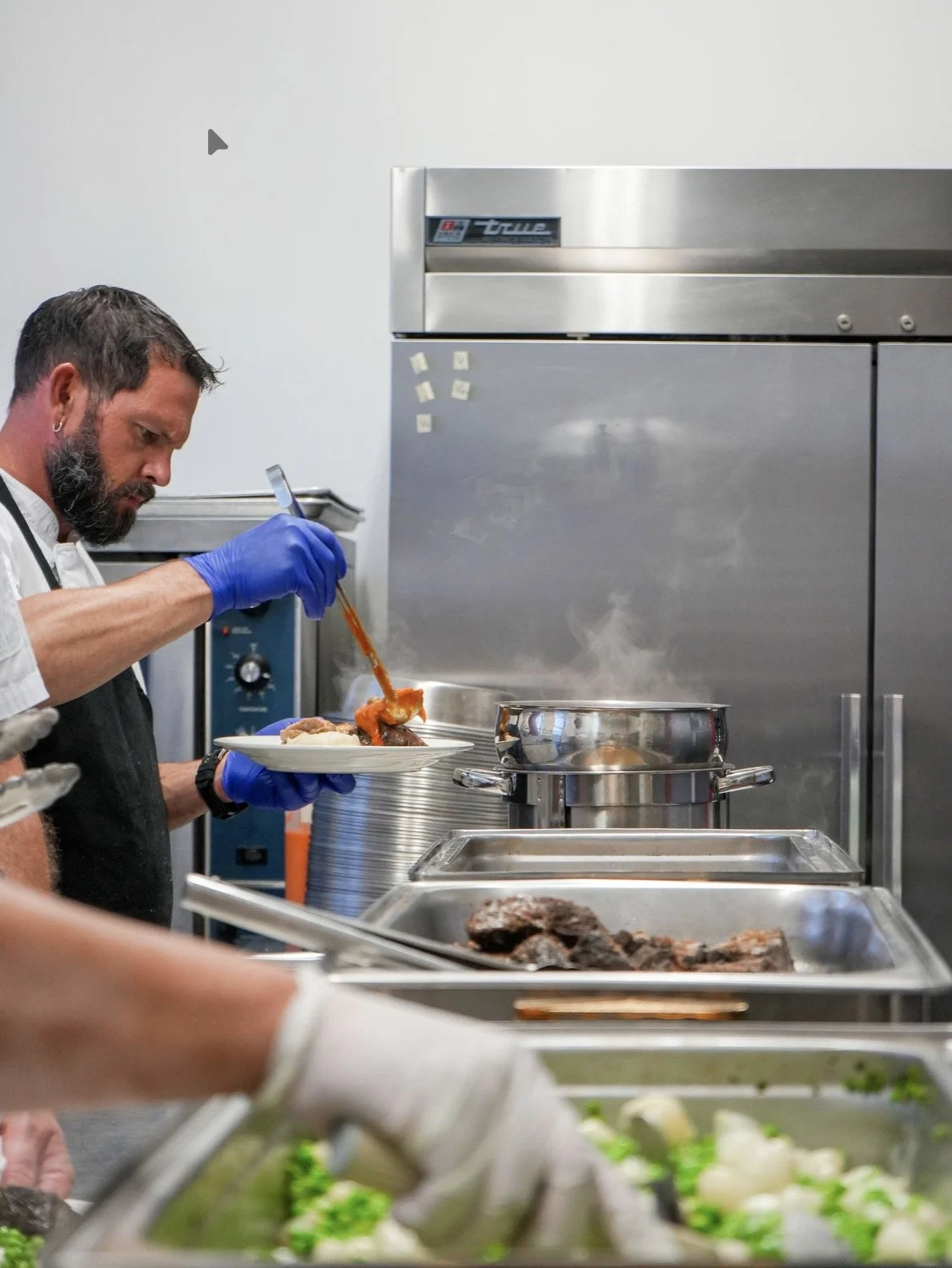 A chef wearing blue gloves serves food on a plate in a professional kitchen with stainless steel appliances and serving trays.