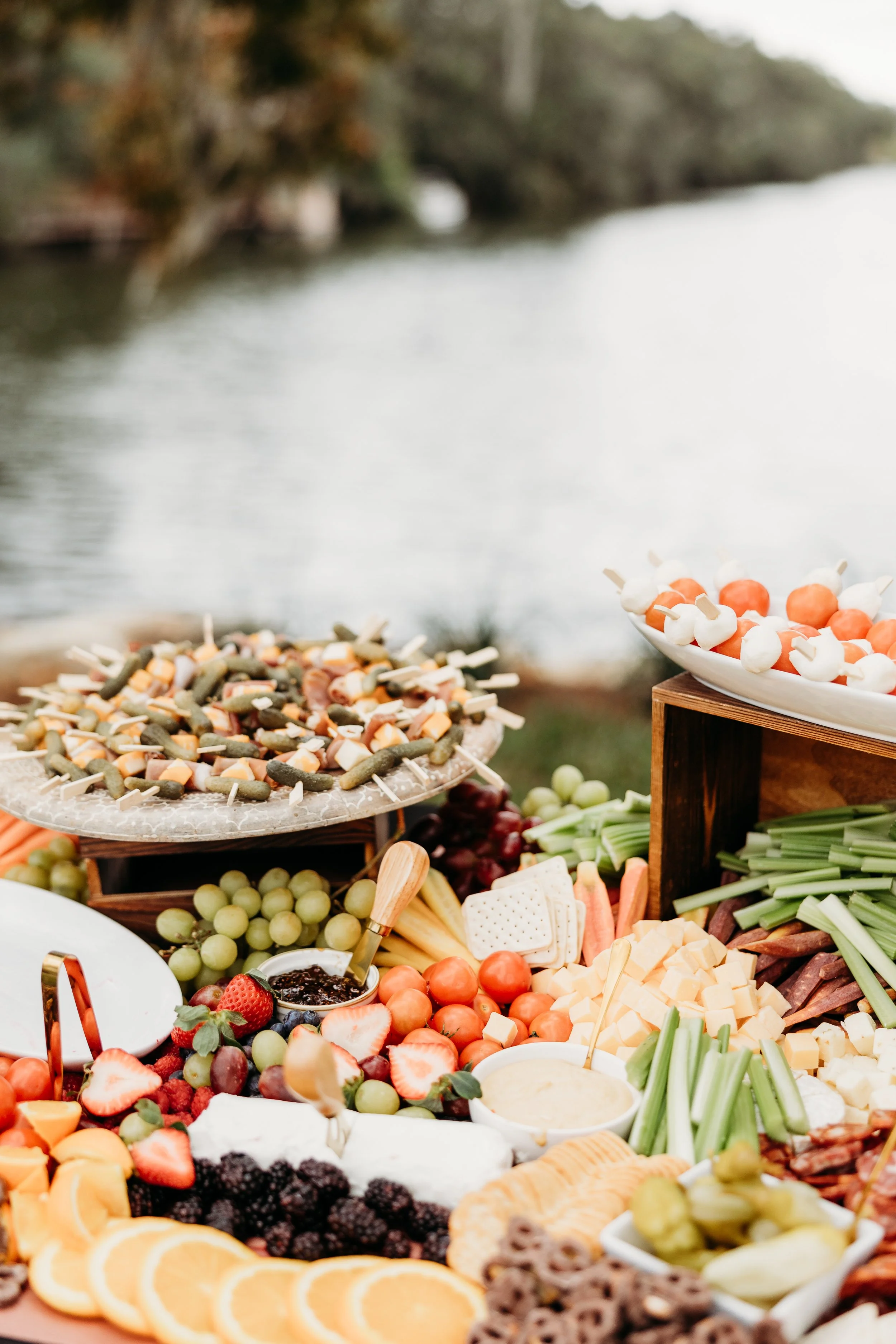 A variety of colorful fruits, vegetables, cheeses, and snacks arranged on a table outdoors near a body of water, with trees in the background.