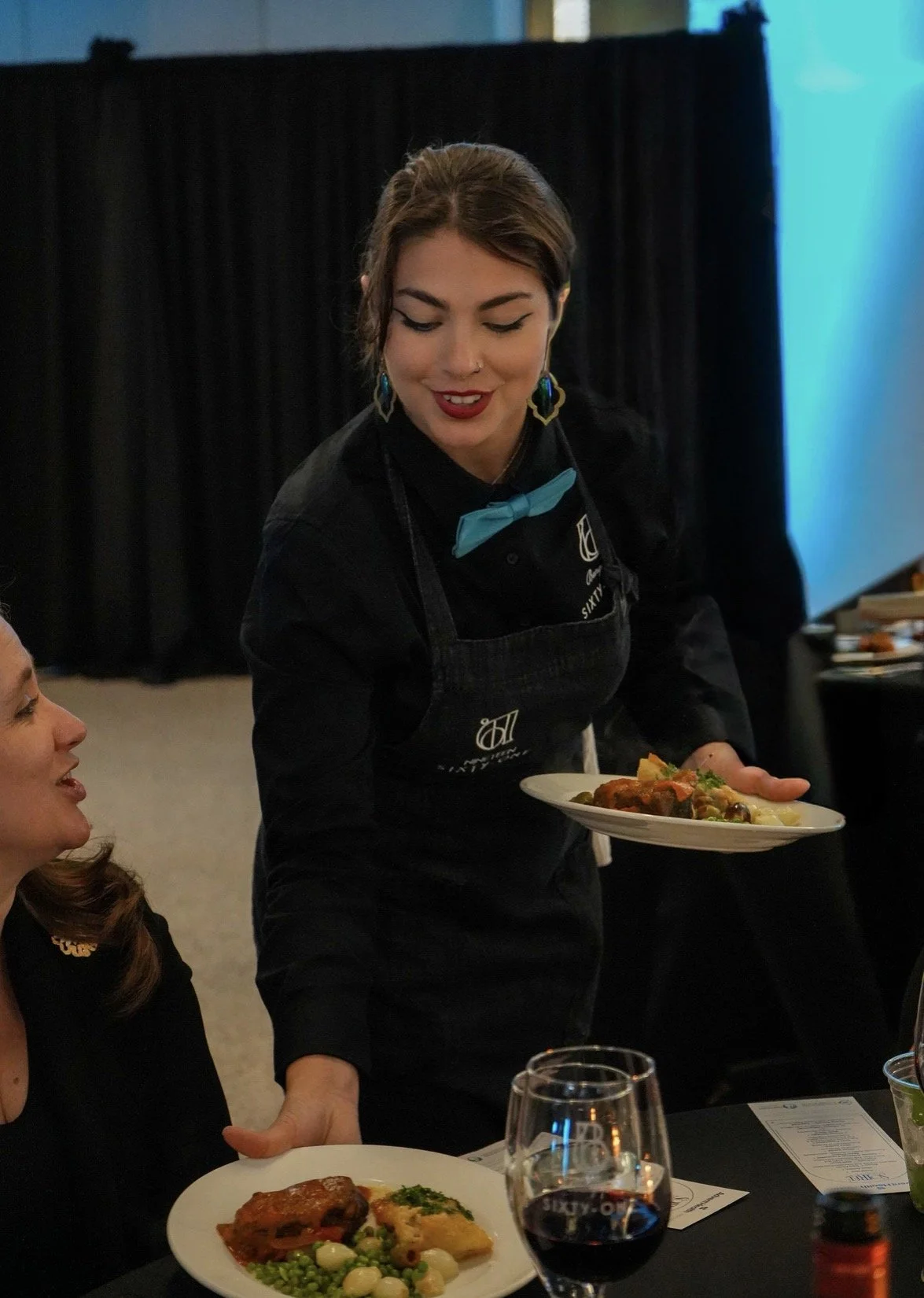 A woman dressed in black, wearing a blue bow tie and colorful earrings, is serving food on a plate at a restaurant or banquet setting. She has short brown hair and is smiling while handing a plate to a seated woman. There are glasses and plates on th