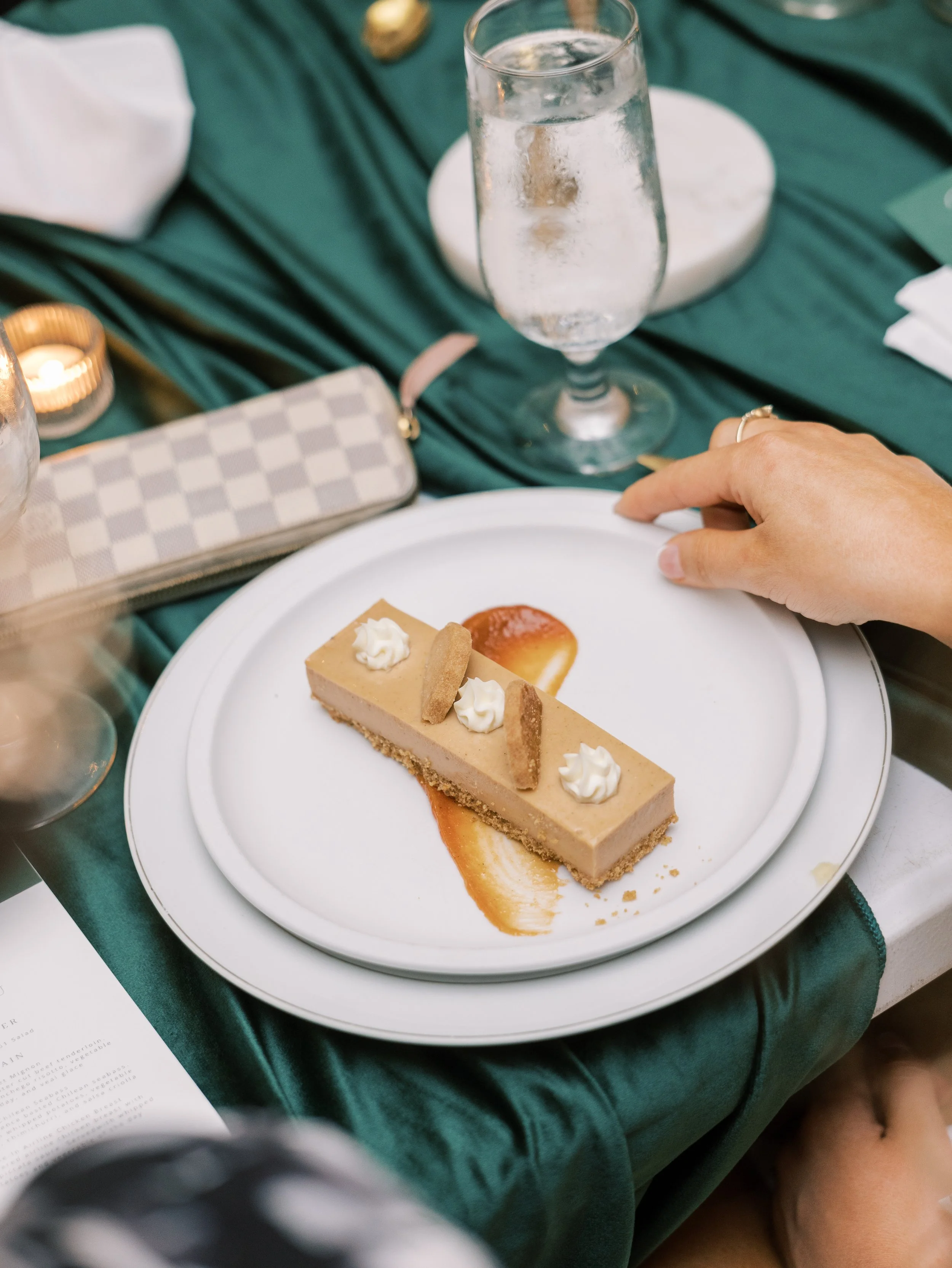 A dessert on a white plate with dollops of whipped cream, caramel sauce, and cookie pieces. A glass of water with ice is in the background on a green tablecloth, with a purse and a candle nearby.
