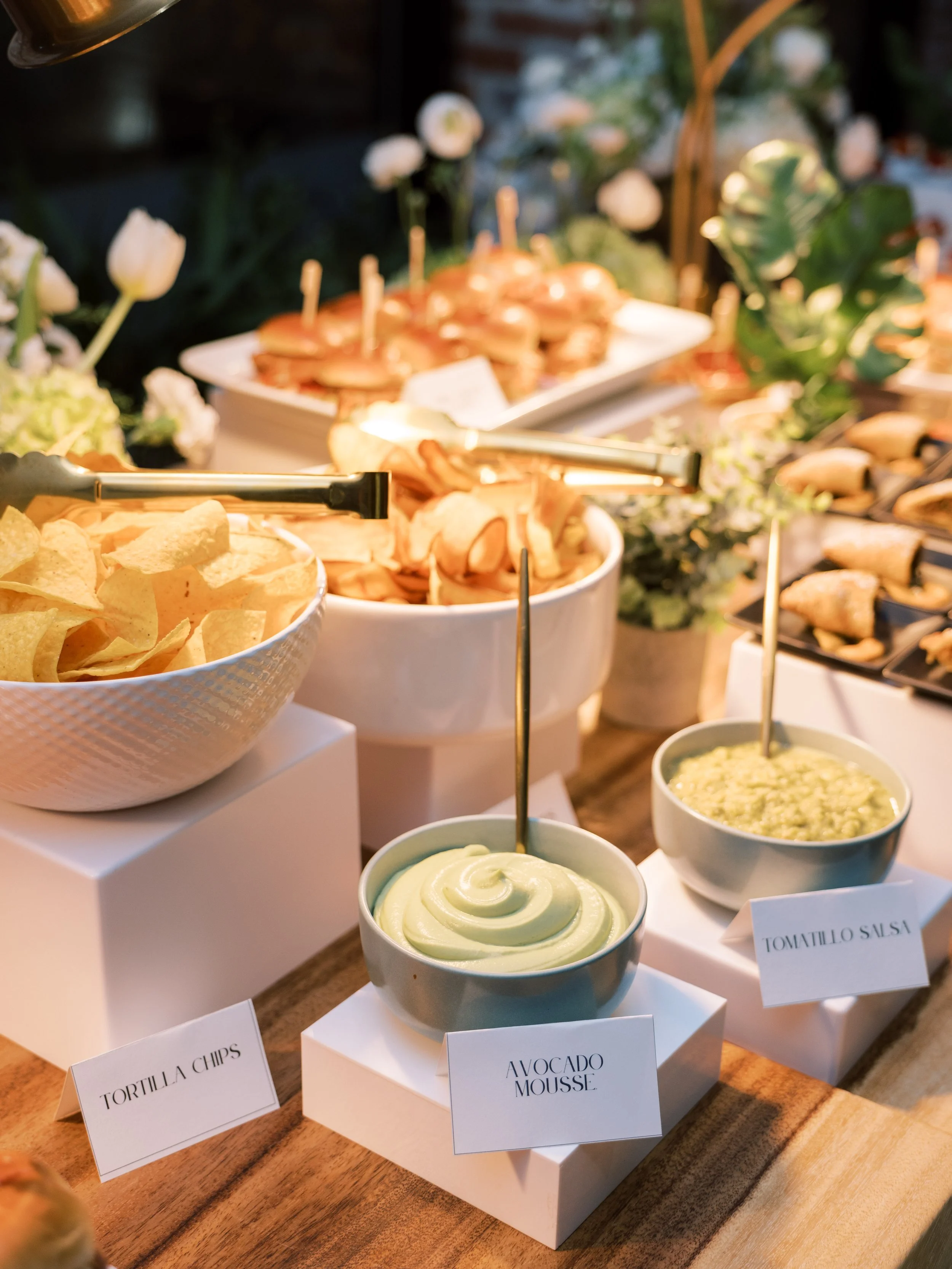 A buffet with bowls of guacamole, salsa, tortilla chips, and small sandwiches, decorated with flowers.