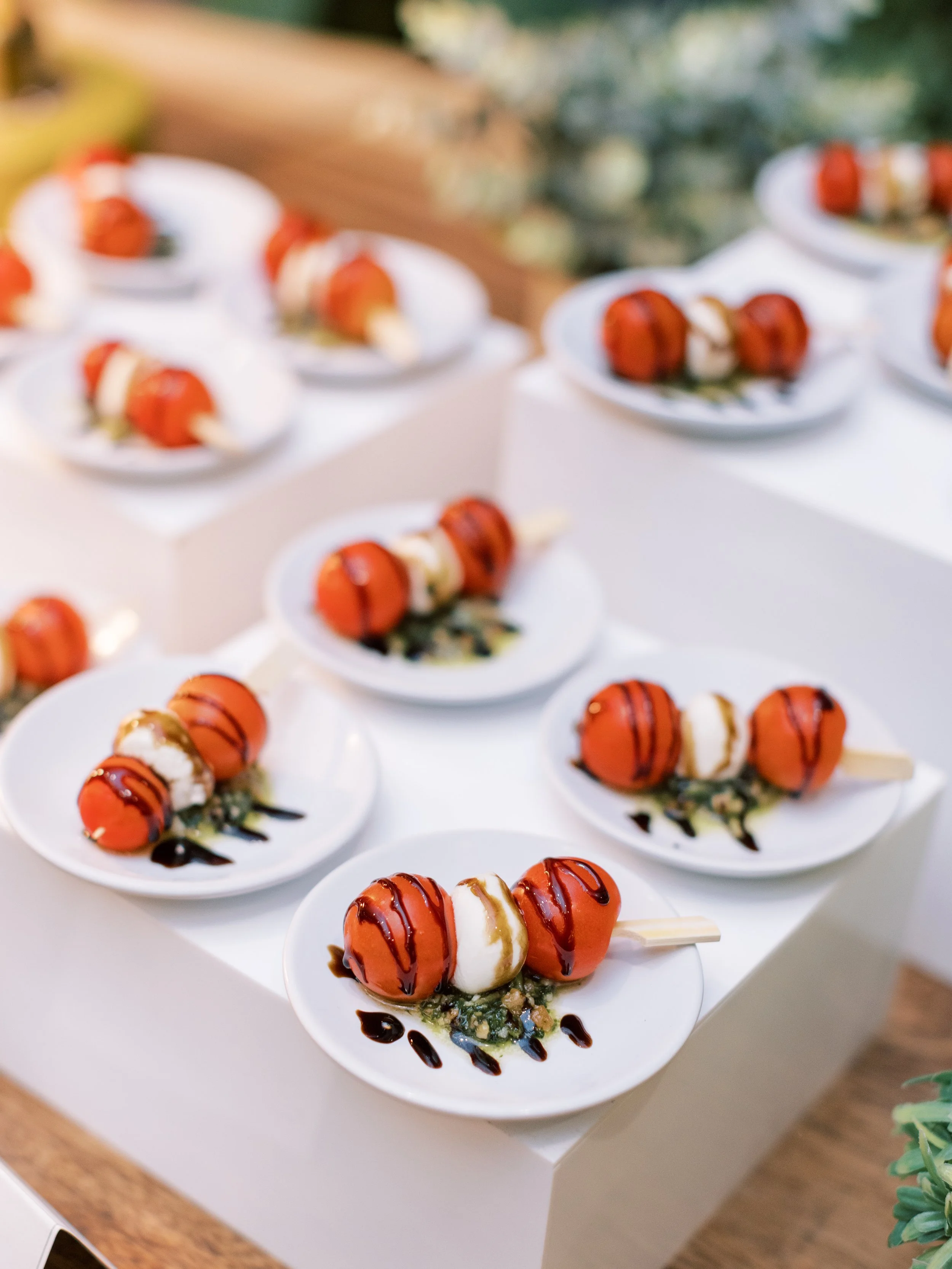 Multiple plates of skewered appetizers with cherry tomatoes, mozzarella cheese, and basil, drizzled with balsamic glaze and placed on a white table.