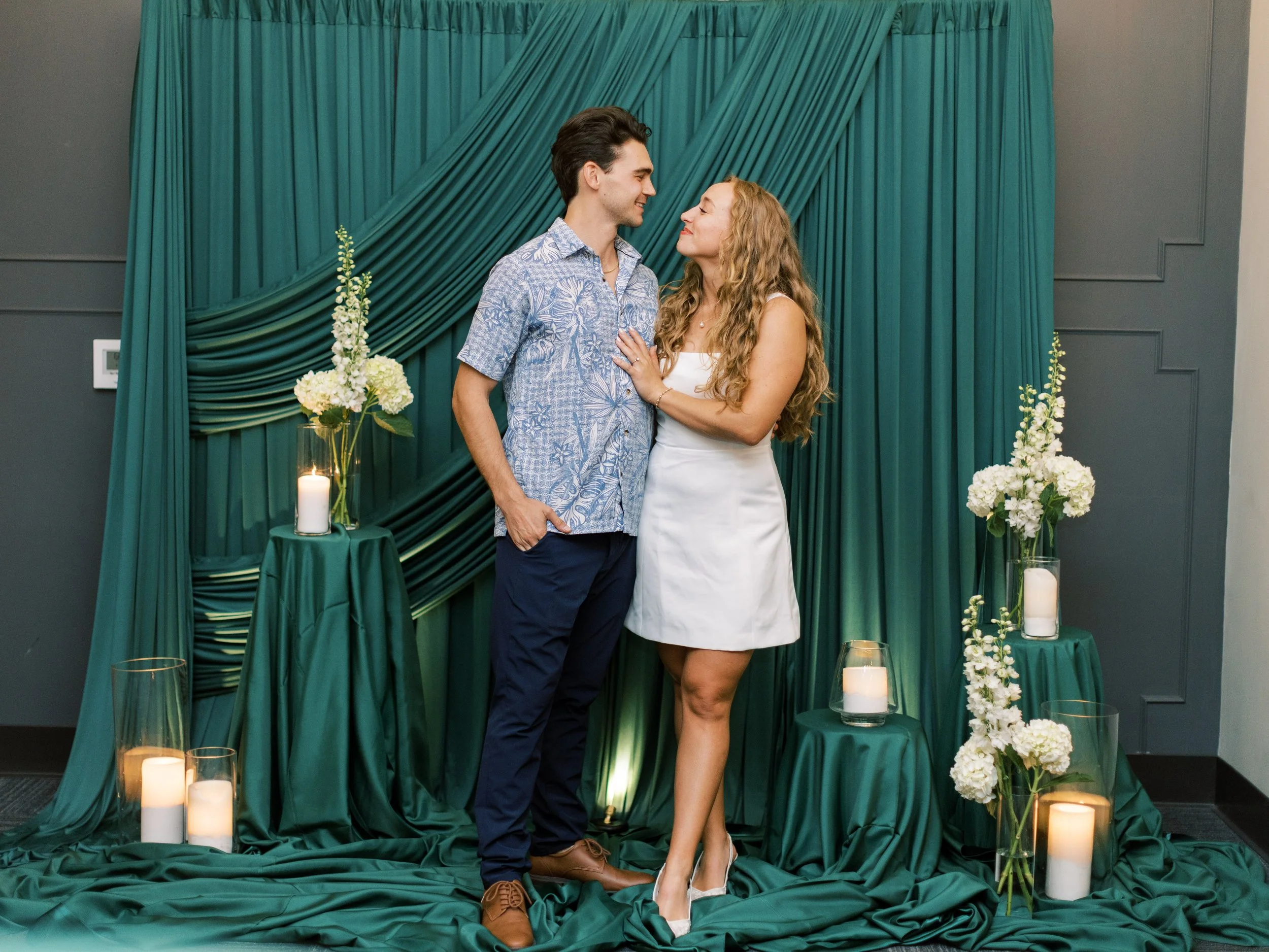 A couple dressed casually and formally getting engaged or married, standing against a teal backdrop with white flowers and candles around them.
