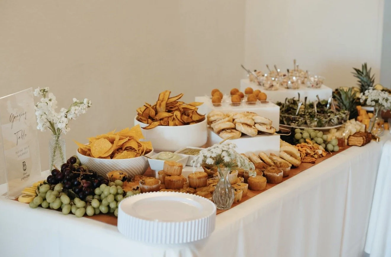 A dessert and snack table with various chips, fruit, cookies, and cupcakes, decorated with flowers and greenery.
