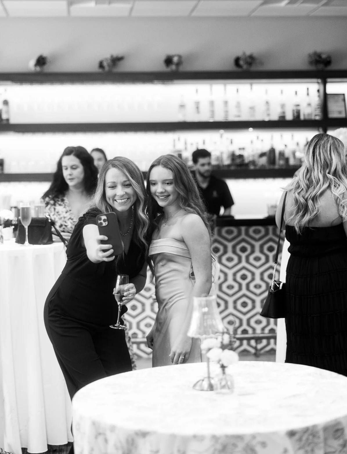 People at a celebration taking a selfie, including a woman and a girl in formal attire, with others in the background, in an indoor event space.