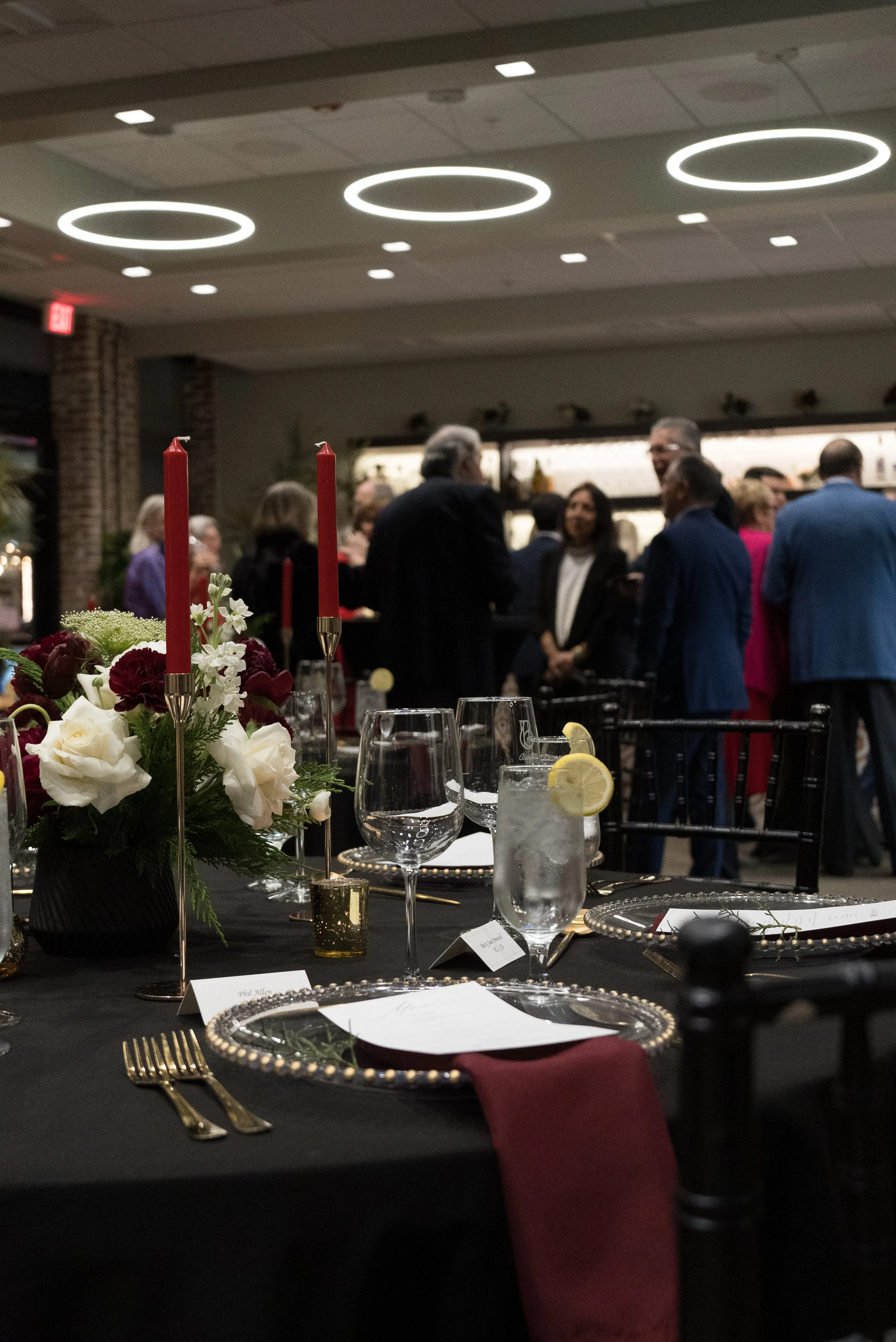 A banquet table set for a formal event with black tablecloth, gold cutlery, glassware, and floral centerpieces, in a decorated indoor venue with people mingling and talking in the background.