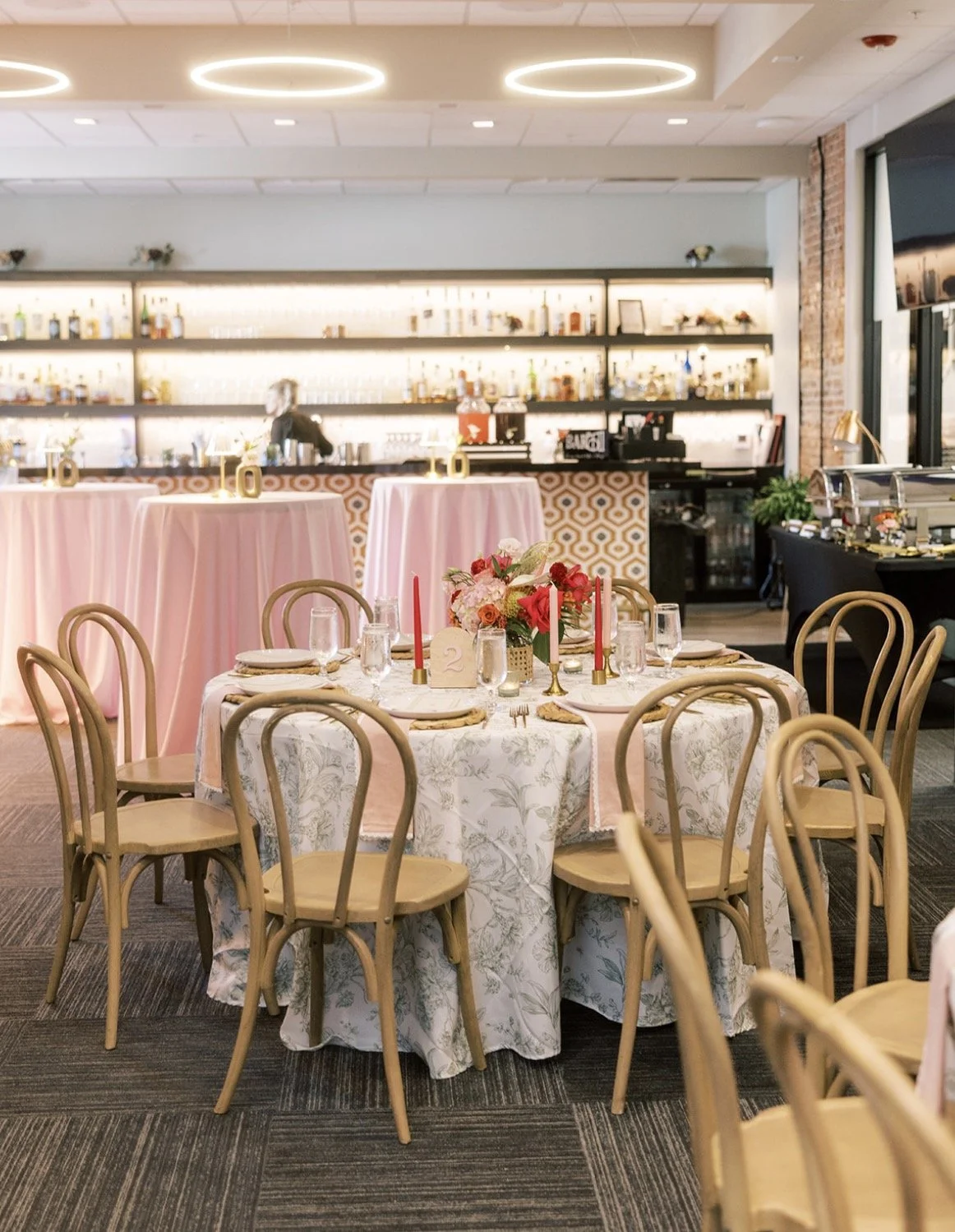 A decorated banquet table with a floral centerpiece, candles, and place settings, set in a restaurant or event venue with a bar in the background and pink table skirts.