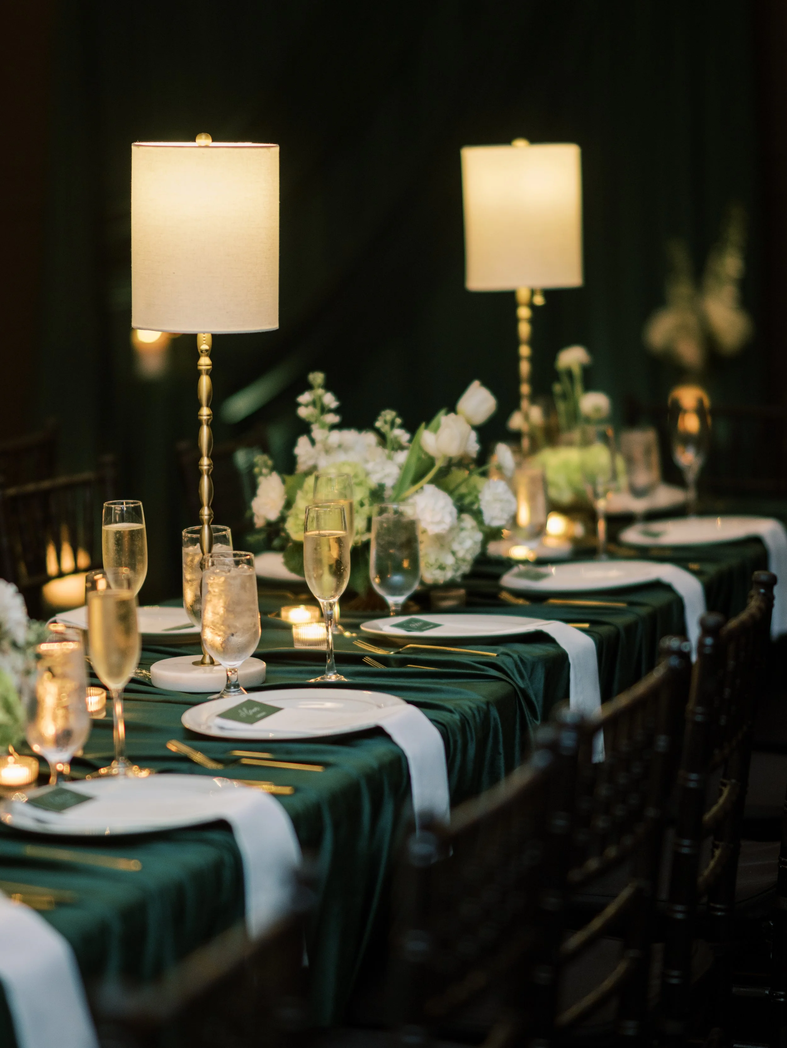 A formal dining table set with white plates, glasses, gold utensils, and white napkins, decorated with floral centerpieces and flanked by lamps with cream-colored shades, in a dimly lit elegant setting.