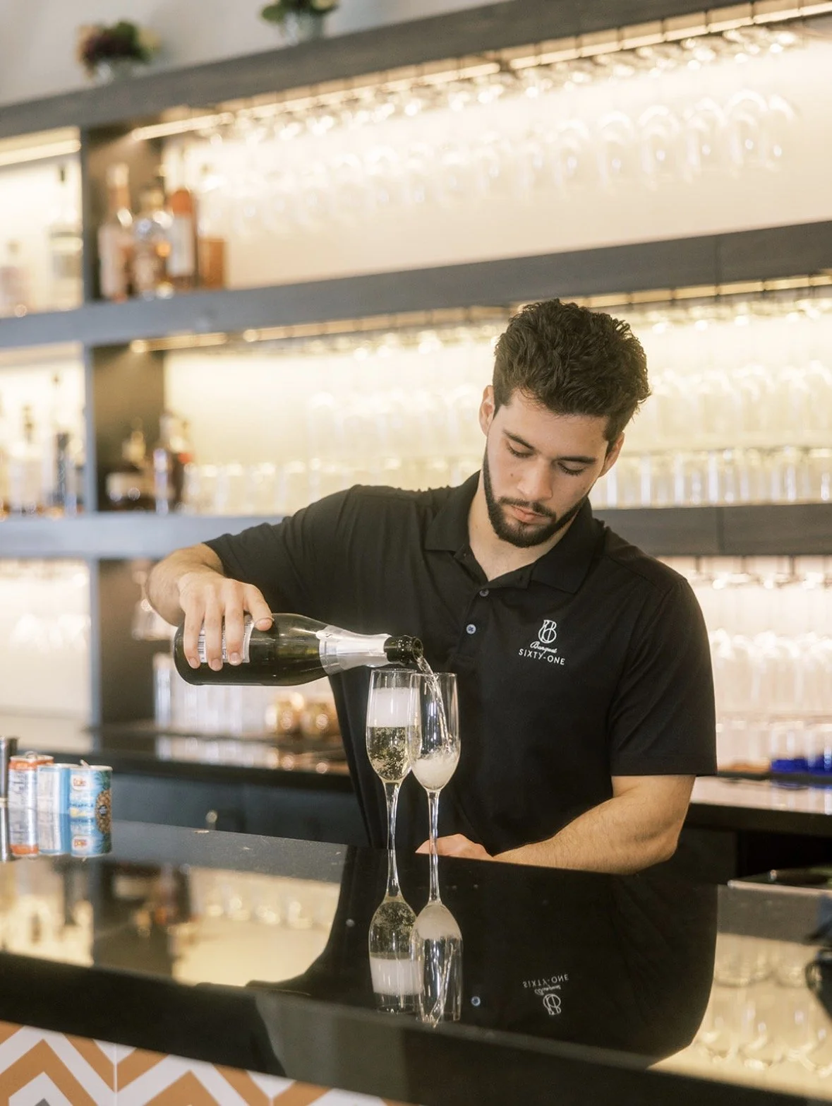 A bartender pouring champagne into two glasses at the bar counter, with shelves of glassware and bottles in the background.