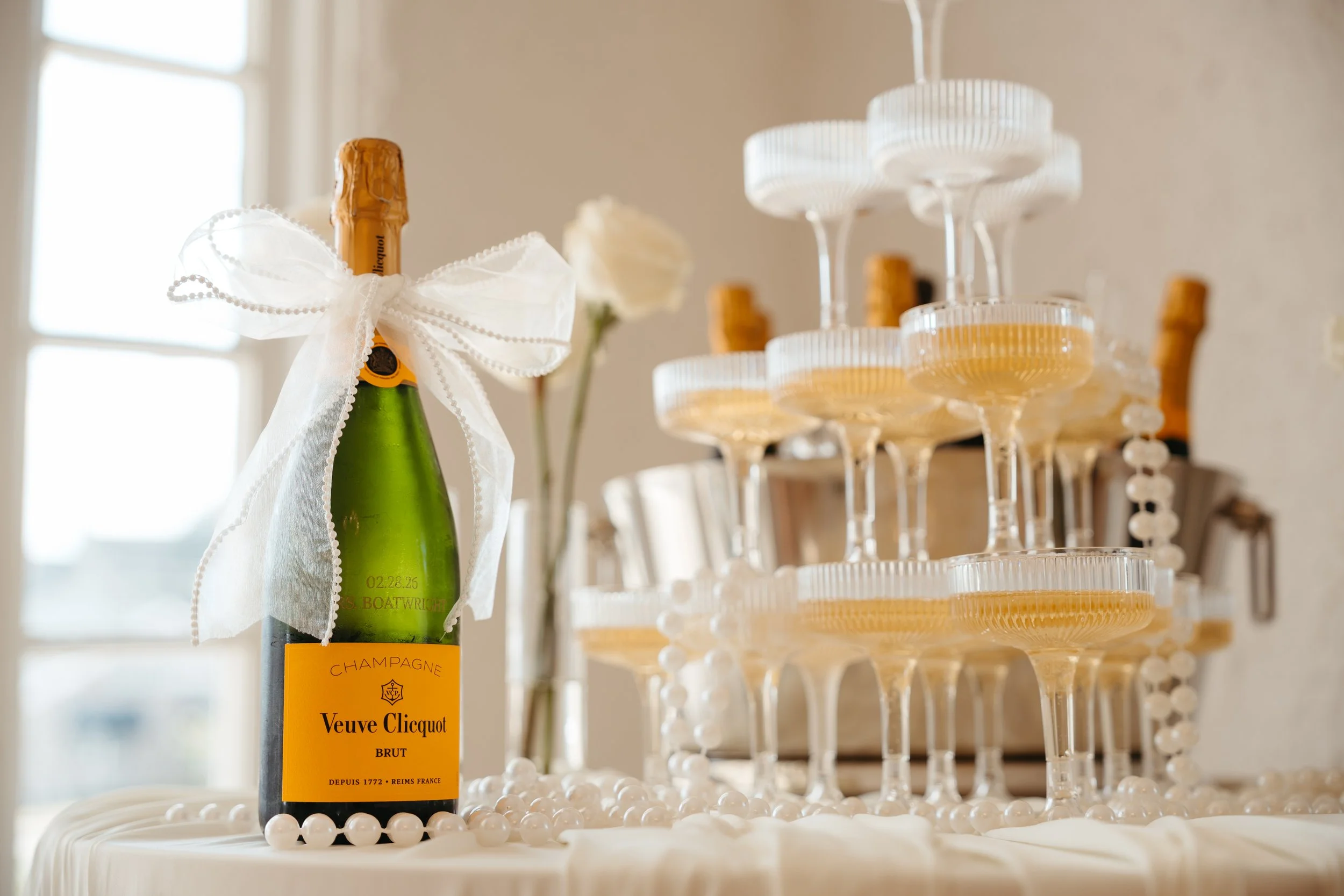 A bottle of Veuve Clicquot champagne with a white ribbon tied around its neck, placed on a decorative table with white beads, and several empty champagne glasses stacked in the background, all inside a bright room with sunlight coming through a windo