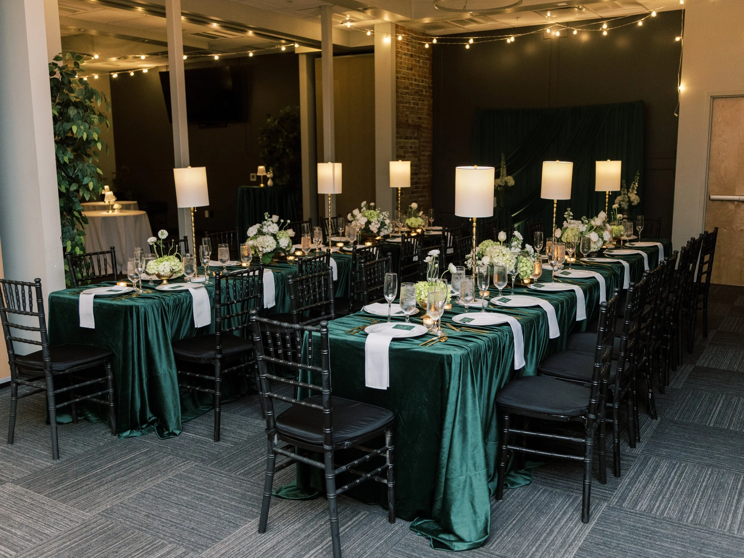 Event space with long tables covered in dark green cloths, decorated with white flowers and set with plates, glasses, and cutlery. Tall white lamps provide lighting, and fairy lights hang from the ceiling. The room has exposed brick and dark walls.