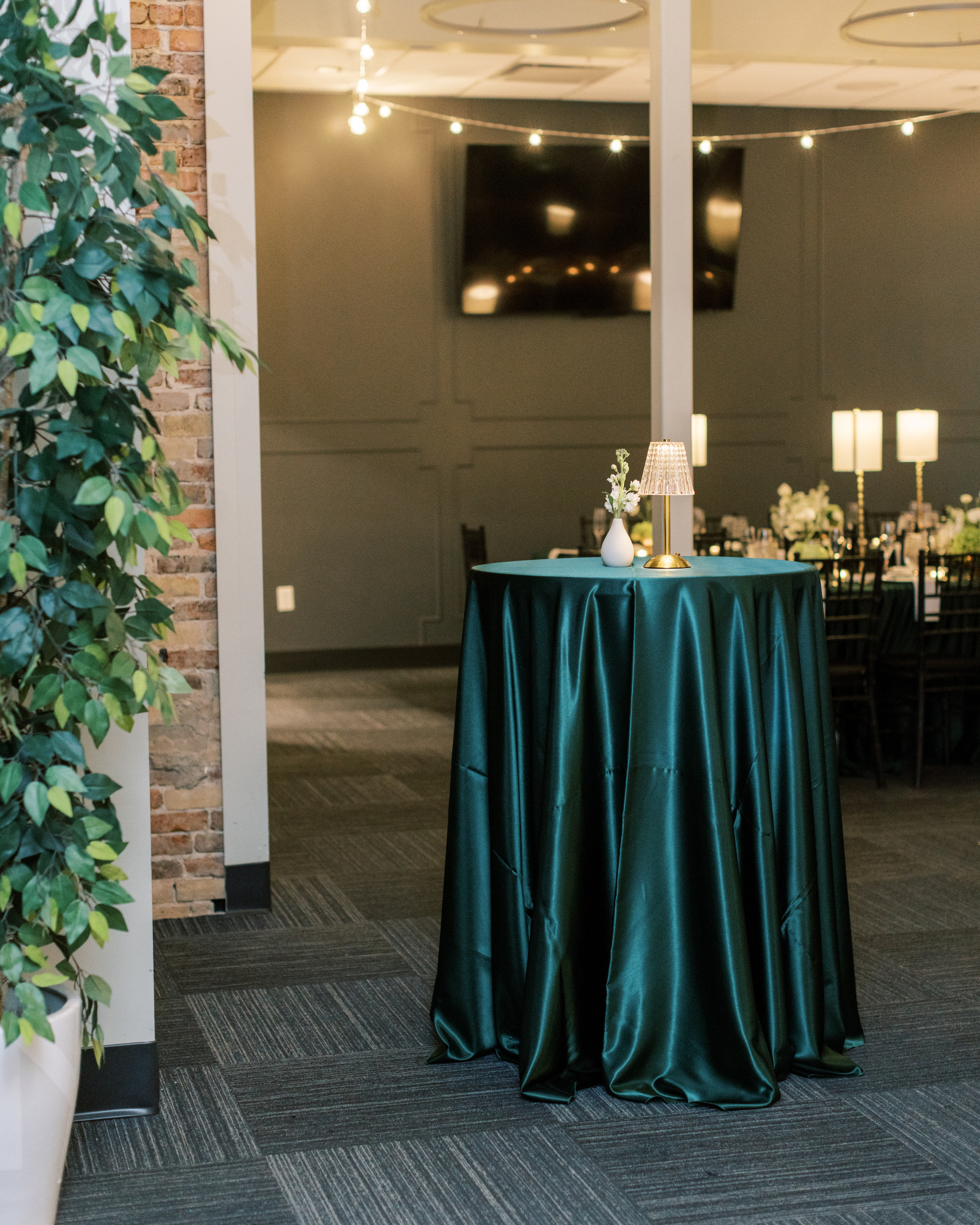 Elegant event space with a round table draped in a dark green satin tablecloth and topped with a gold and white lamp and a white vase with flowers. The background features string lights hanging from the ceiling, a large television on the wall, and ta