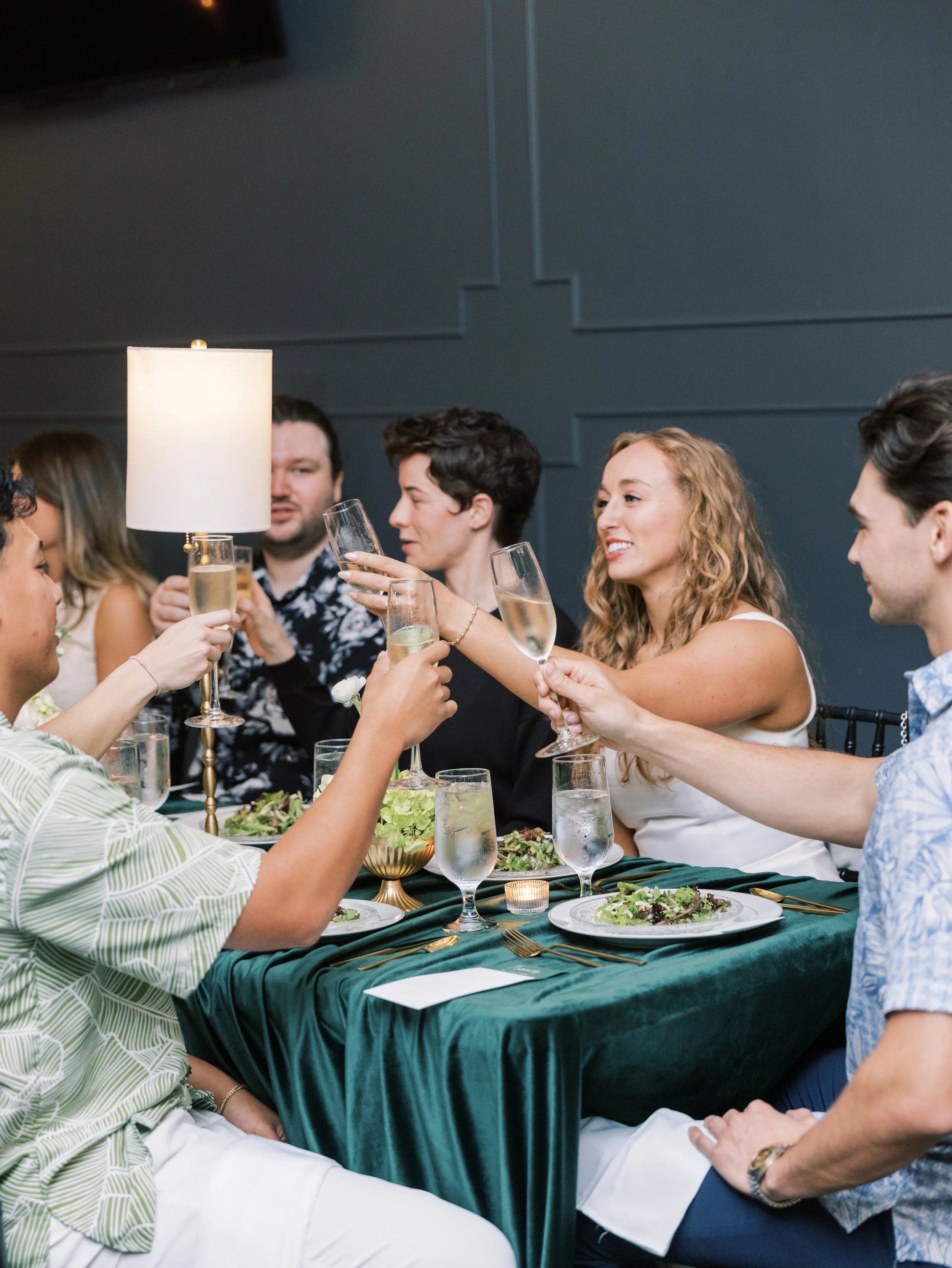 A group of people at a dinner party raising champagne glasses for a toast, with a dark blue wall and a table set with plates, salads, a candle, and a table lamp in the background.
