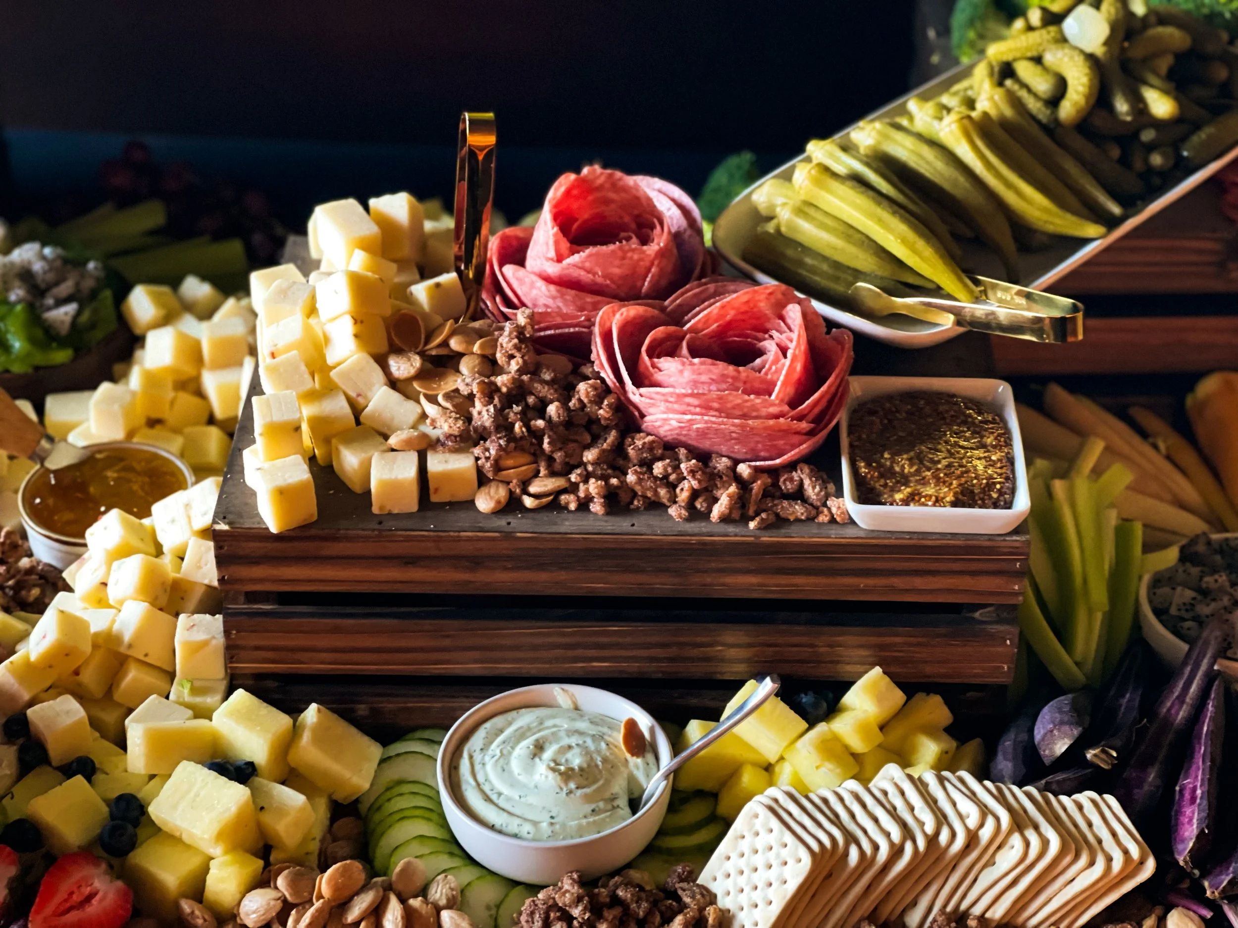 A large cheese platter with various cheeses, meats, crackers, fruits, nuts, and dips arranged on a wooden table, with tongs and bowls of condiments.