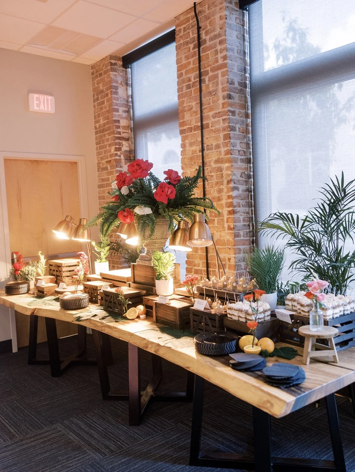 A wooden table with floral decorations, cakes, and dessert items set up against a brick wall with large windows. There are several potted plants and metallic lamps illuminating the table.