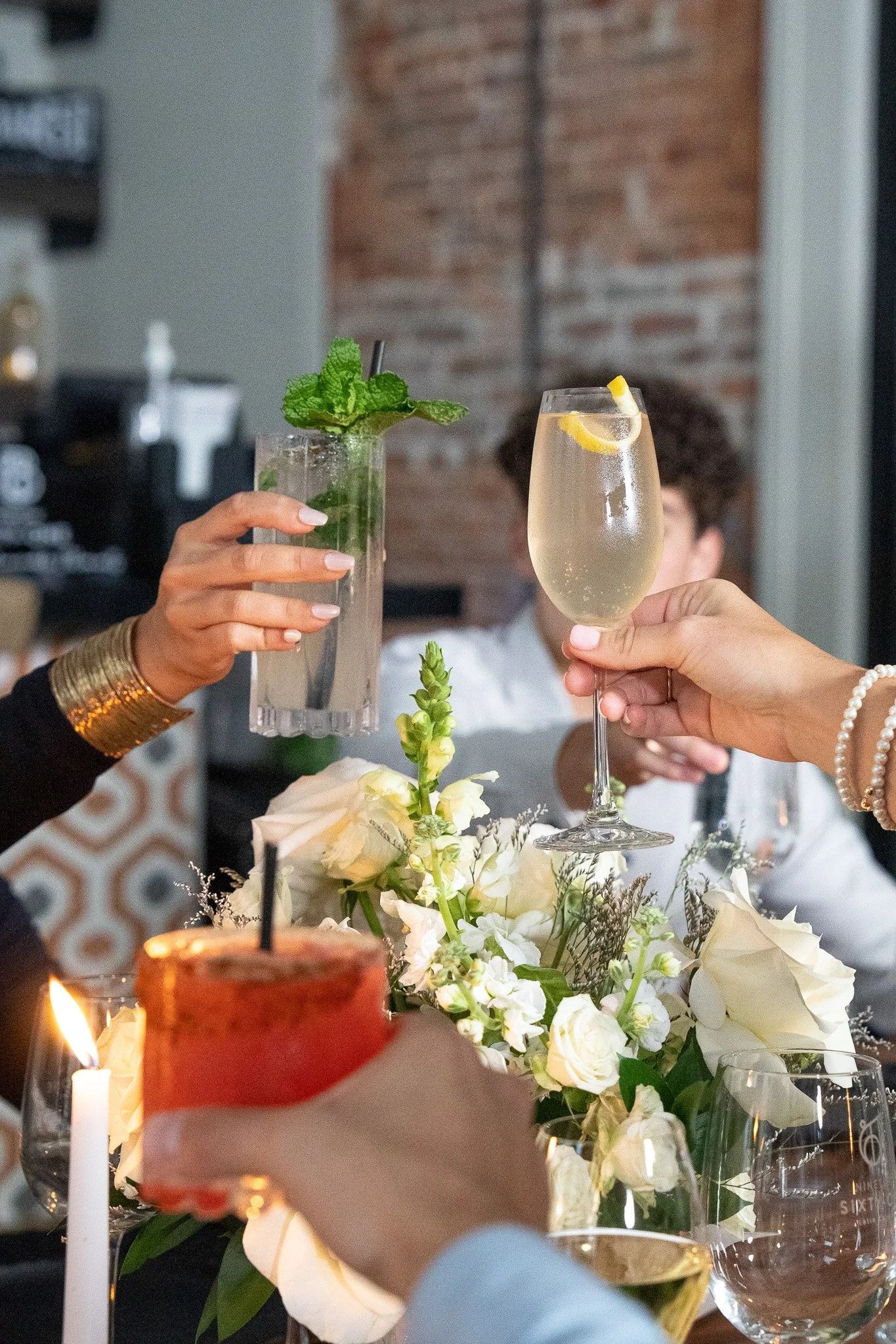 People raising glasses of champagne and mint lemonade in a toast at a celebration with floral arrangements, candles, and a brick wall in the background.