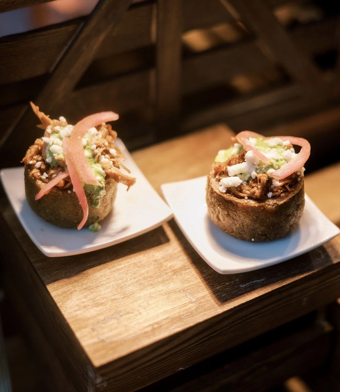 Two Mexican dish bowls called 'tortillas rellenas' with different fillings, topped with pickled onions and green sauce, served on small white plates on a wooden table.