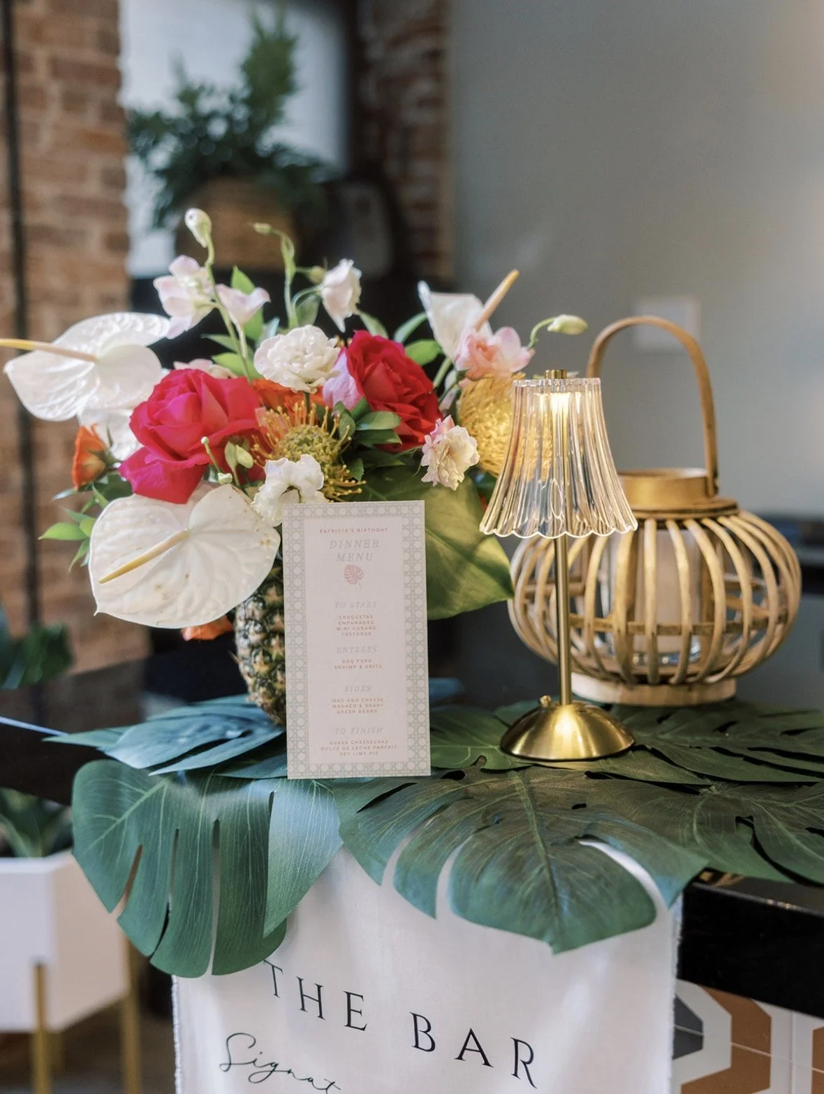 A floral arrangement with pink and white flowers on a table with a large green leaf placemat, a small golden table lamp with a pleated shade, and a wicker lantern. There is a small menu card in front of the flowers, and a branded tablecloth with the 