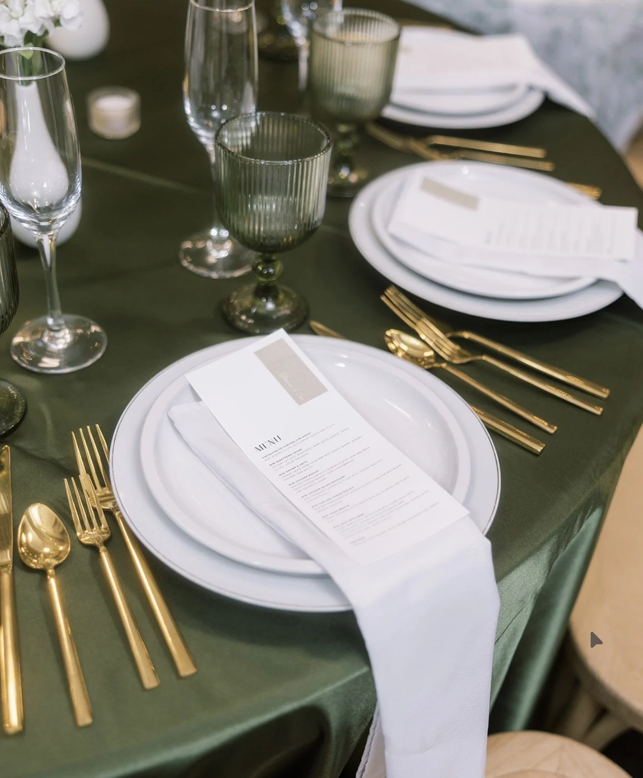 A formal table setting with gold flatware, white plates with cutlery, green glassware, and a menu on a white napkin, set on a dark green tablecloth.