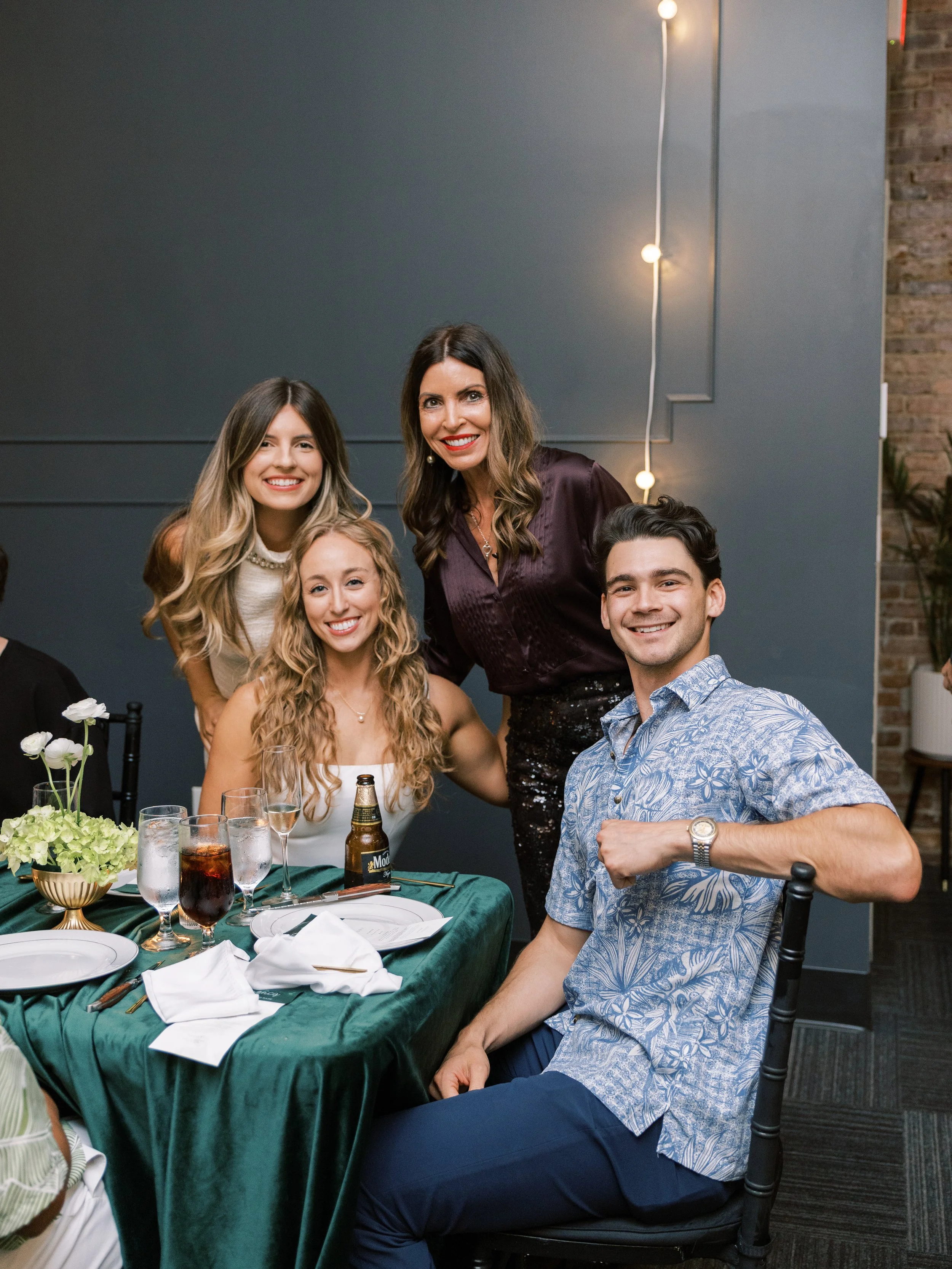A group of five people gathered around a dining table at a formal or semi-formal event. The table is set with plates, glasses, and silverware, and has flowers as decoration. The background features a dark wall with string lights and a small section o