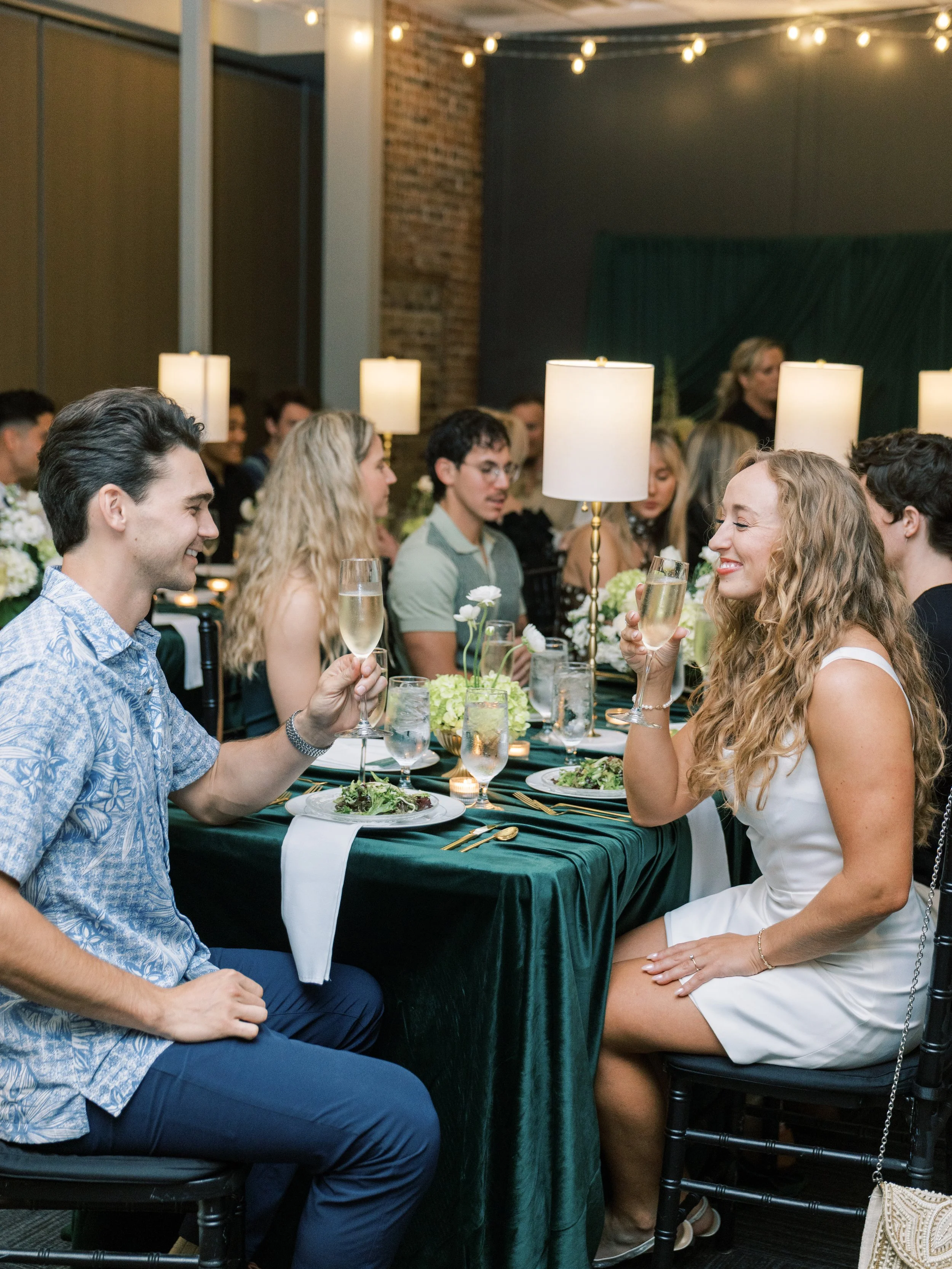 A group of people at a formal dinner party, with men and women sitting at a table with green tablecloths, white flowers, and candles. Many are holding glasses of champagne and smiling.