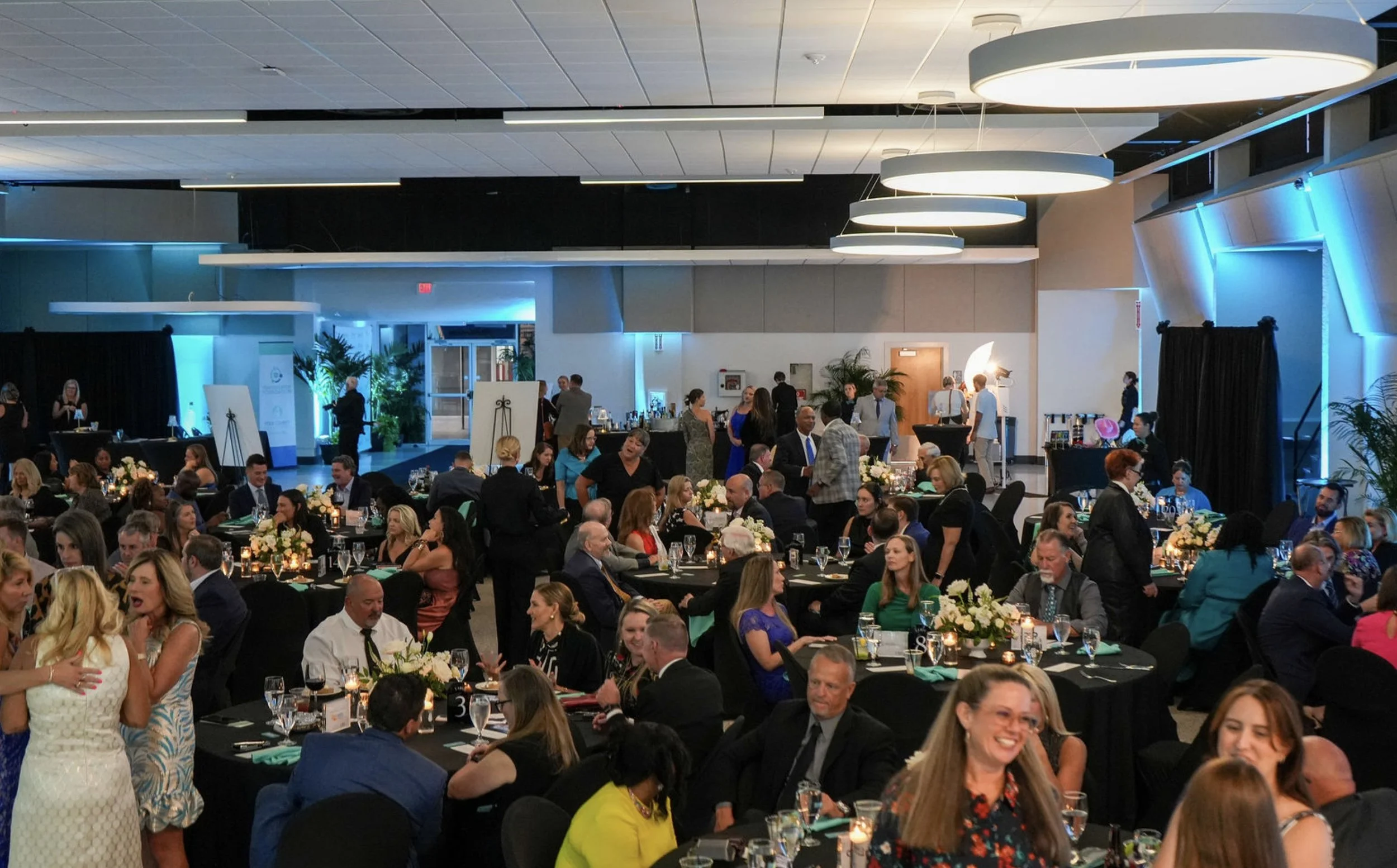 A large banquet hall filled with many round tables occupied by well-dressed guests attending a formal event. There are floral centerpieces, candles, and glassware on the tables. In the background, people are standing by a bar and engaging in conversations. The hall has modern lighting fixtures and blue accent lights.