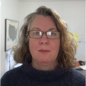 A woman with curly hair wearing glasses and a dark sweater, standing indoors in front of a window and framed art.