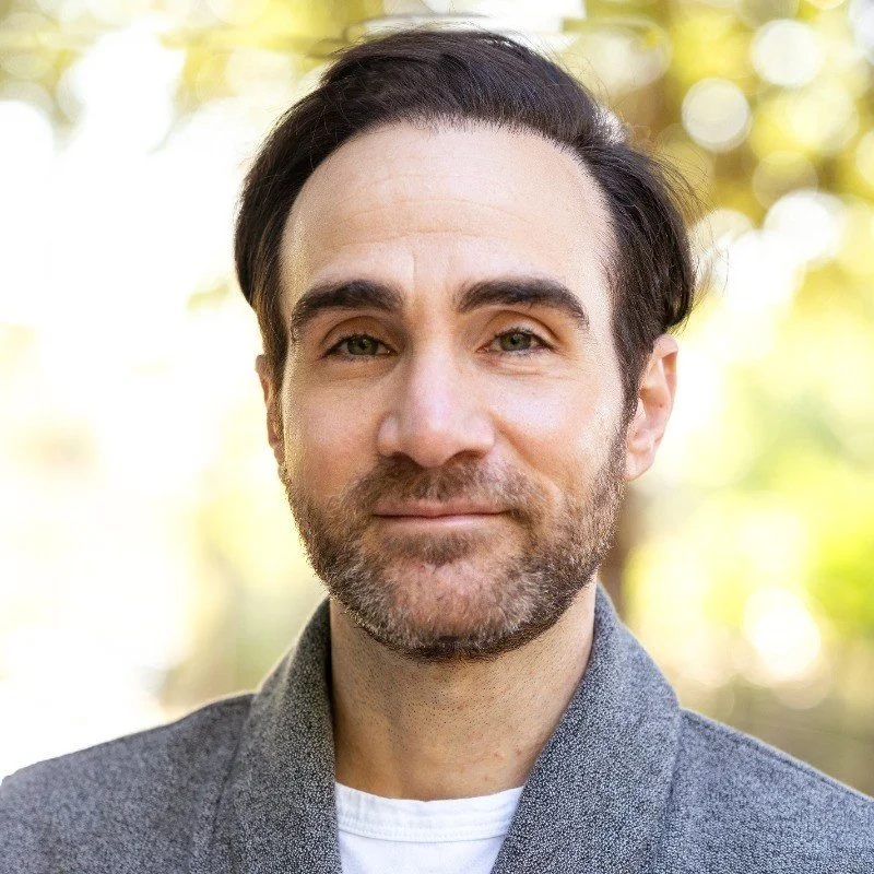 A man with dark hair and a beard smiling outdoors with a blurred background of yellow and green foliage.