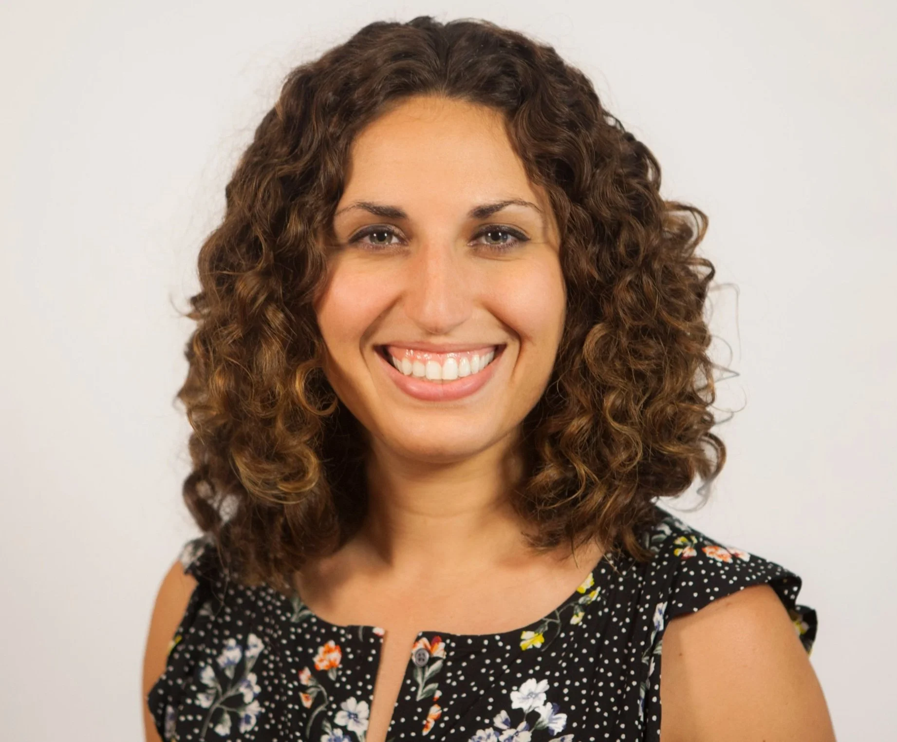 A woman with curly brown hair wearing a black floral patterned top smiling at the camera against a plain white background.