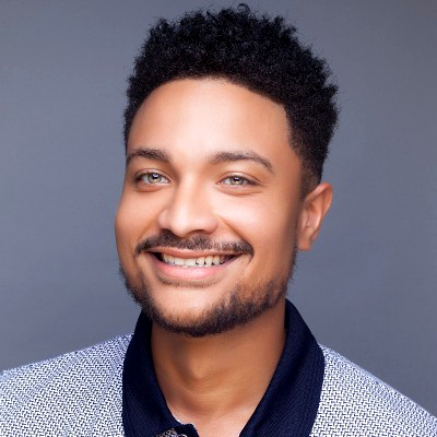 Smiling young man with curly hair and a beard, wearing a blue and white patterned shirt, against a gray background.