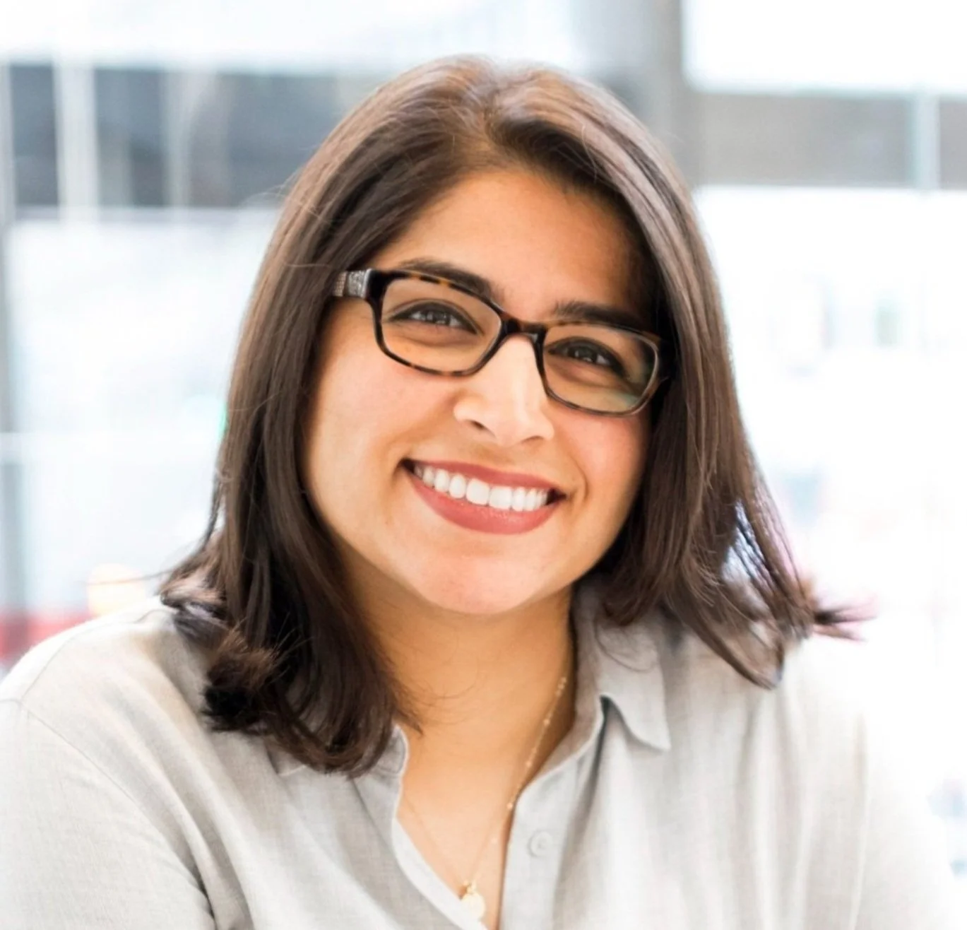 A smiling woman with shoulder-length brown hair, wearing glasses and a light-colored shirt, sitting in a modern office setting.