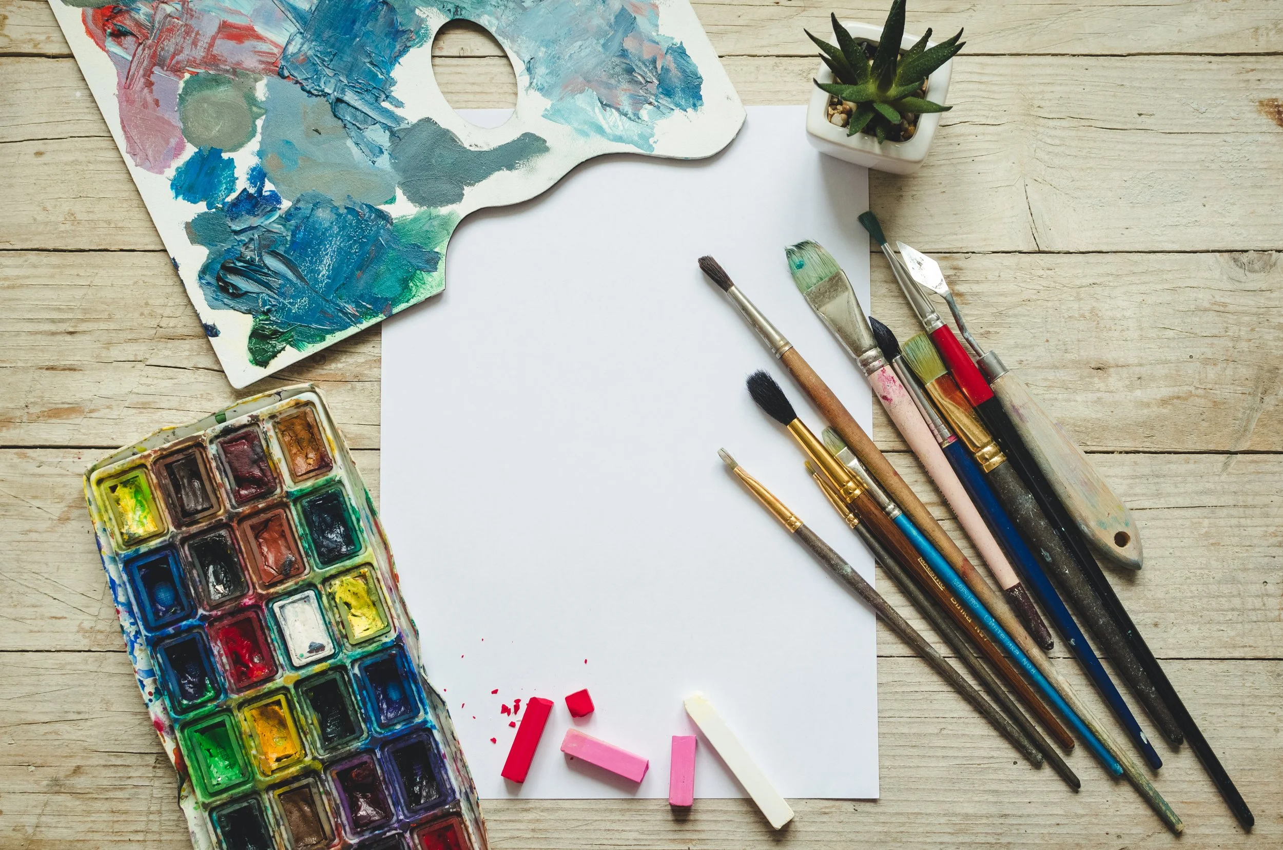 An artist's workspace with a paint palette, paintbrushes, watercolor set, blank sheet of paper, a small potted succulent, and colorful chalk pieces on a wooden surface.