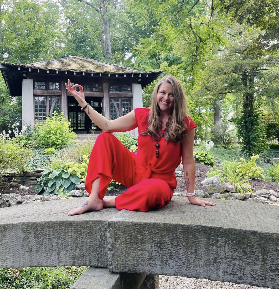 A woman in a red outfit sitting cross-legged on a stone bench in a lush, green outdoor garden, smiling and making an 'okay' gesture with her right hand, with a traditional-style wooden building in the background.