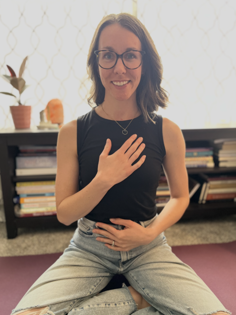 A woman with shoulder-length brown hair, glasses, and a black sleeveless top sitting cross-legged indoors on a purple yoga mat, with a hand on her stomach and the other over her heart, smiling at the camera.