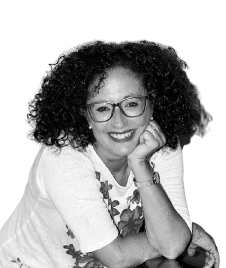 Donna con capelli ricci e occhiali, sorridente, che indossa una maglietta bianca con motivo floreale.