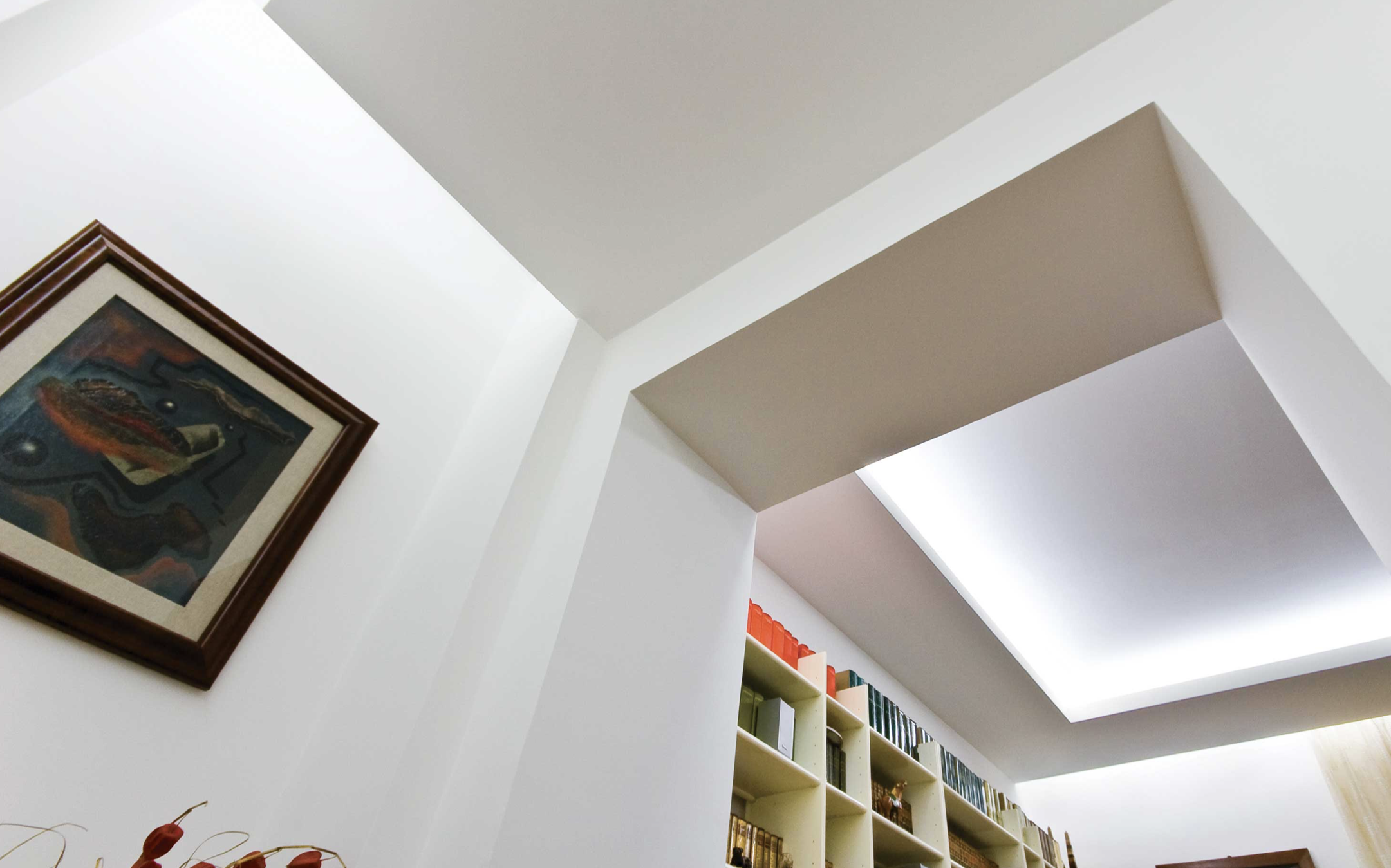 Interno di una stanza con pareti bianche, una libreria piena di libri e una cornice con un quadro astratto sulla parete.