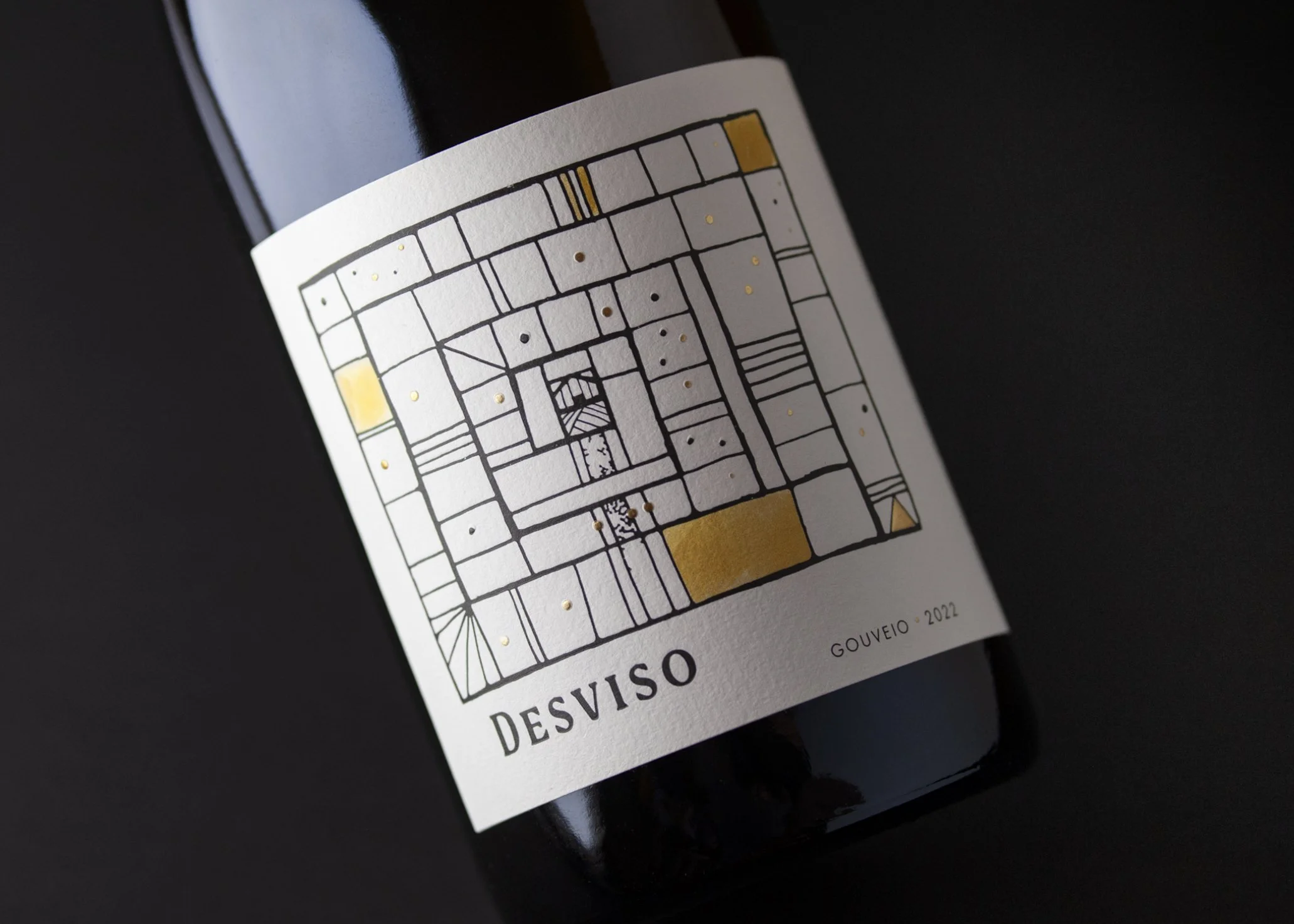 Close-up of a dark glass bottle with a white label featuring a black and gold abstract geometric design, the word "DESVISO" and the year "2022" printed at the bottom.