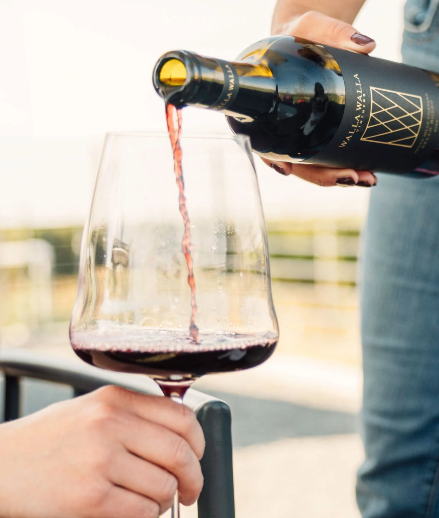 A person is pouring red wine from a dark wine bottle into a glass outdoors during sunset.