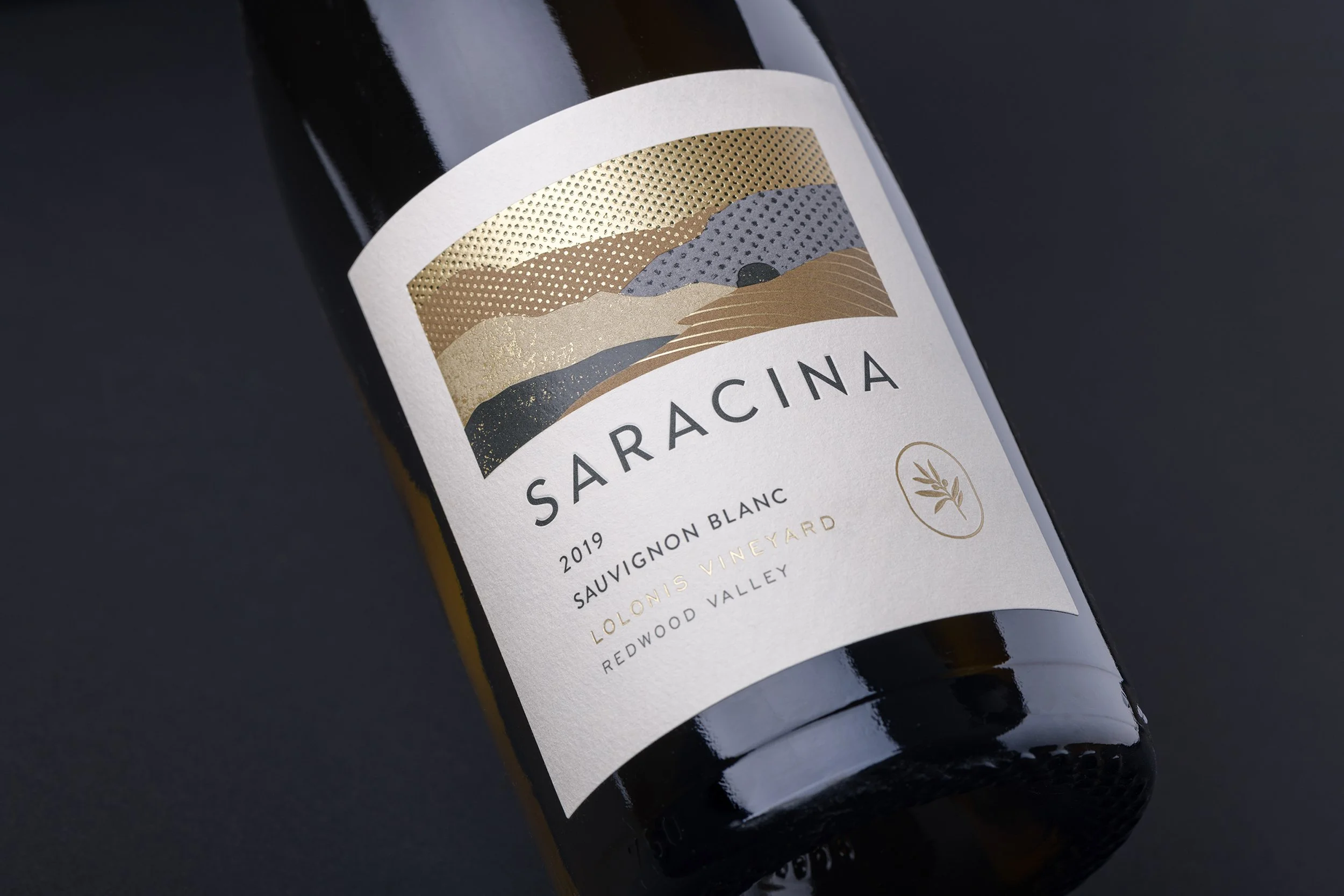 Close-up of a wine bottle label with abstract gold, gray, and beige mountain landscape design, and the text "SARACINA" and details about the wine.