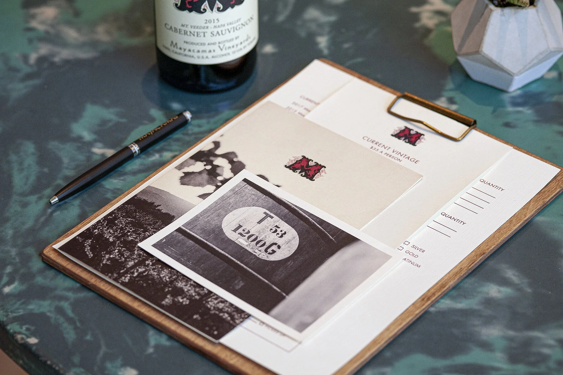 A presentation board with a sample of a whiskey label, a piece of paper with the text "1200G," a pen, a wine bottle, and a small geometric object on a green marble table.