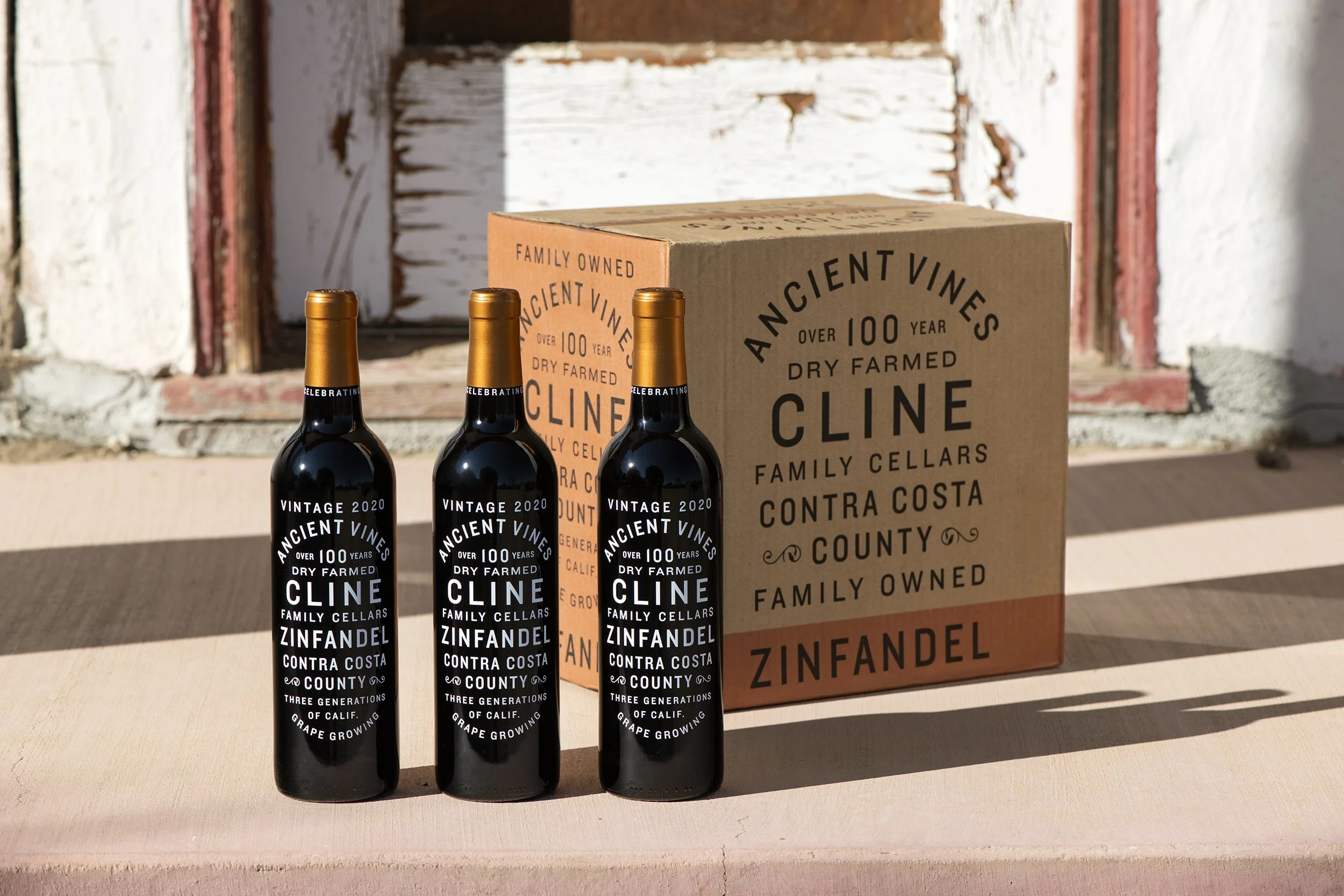 Four bottles of Cline Zinfandel wine with black labels and gold caps, placed in front of a cardboard box from Cline Family Cellars, on a wooden surface against a rustic, weathered background.