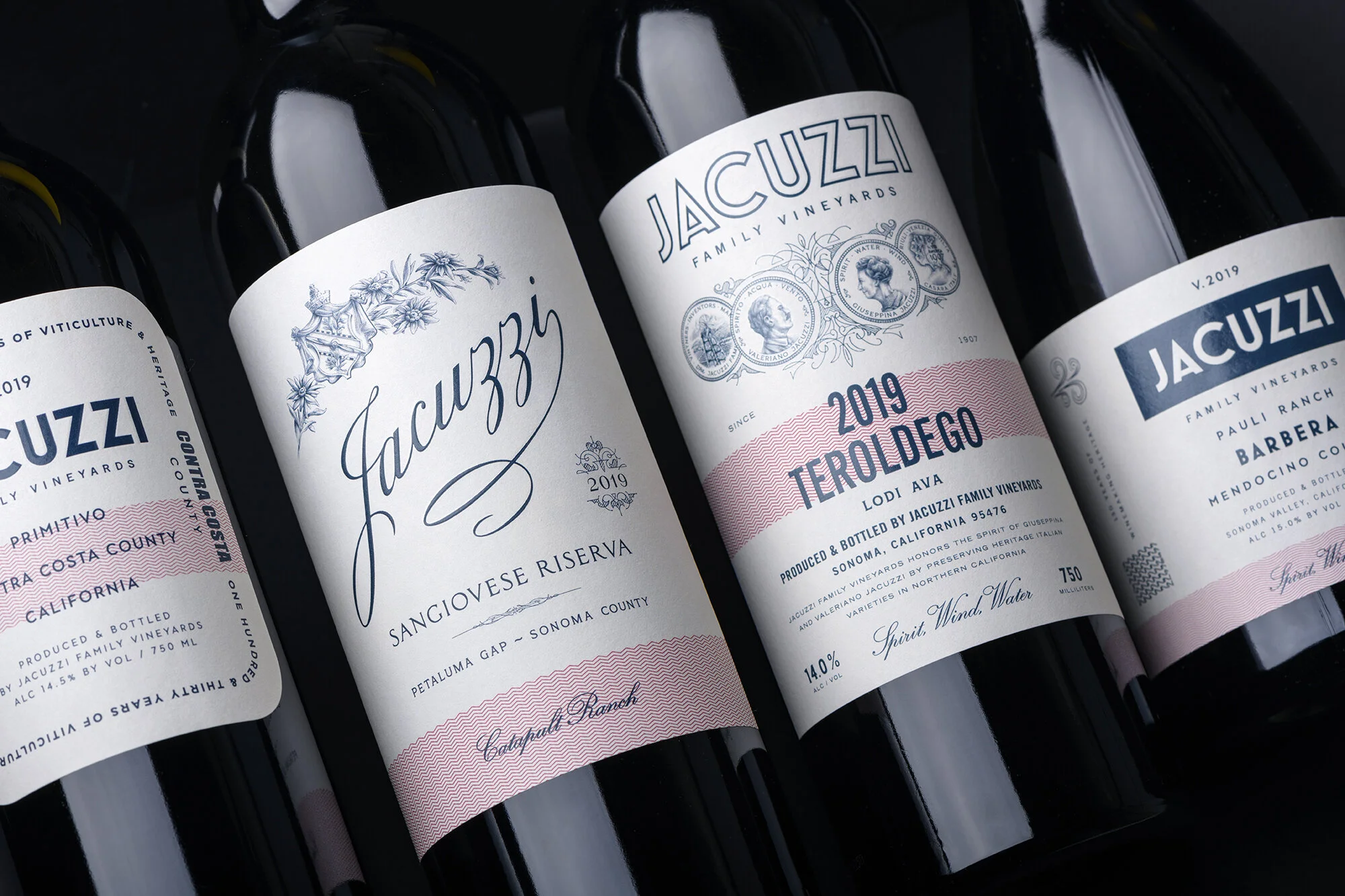 Close-up of wine bottles with labels from Jacuzzi Family Vineyards, including a bottle of Sangiovese Riserva and a Teroldego from 2019, set against a dark background.