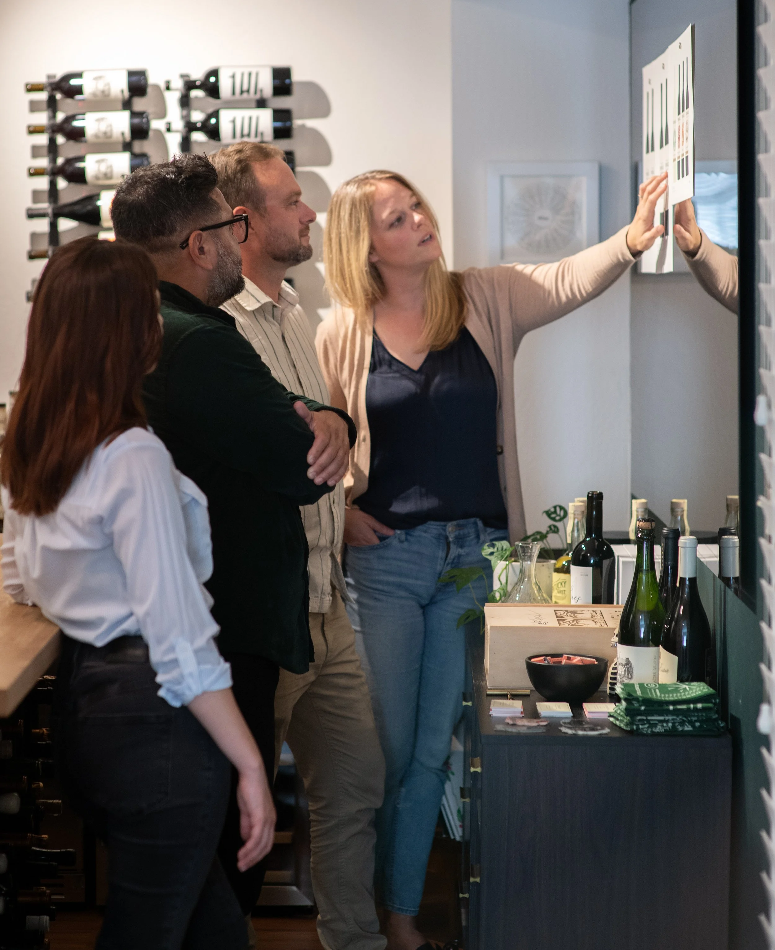 A group of four people, two men and two women, tasting or selecting wine in a wine shop or tasting room. They are standing in front of a display of wine bottles and a tasting table with bottles and glasses.