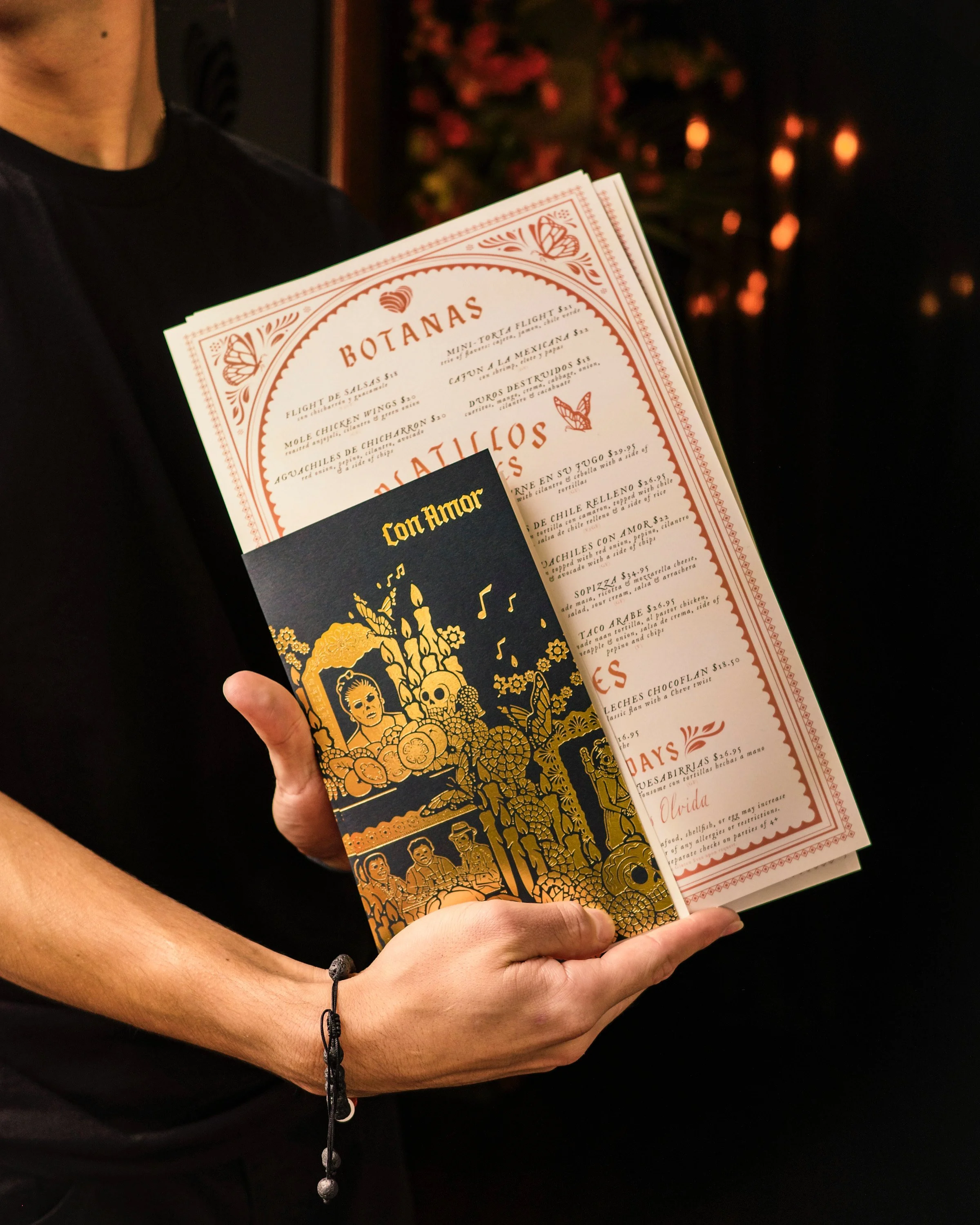 A person in a black shirt holding a menu with floral and skull artwork, with a dark background and out-of-focus lights or candles in the distance. Con Amor—Napa, California