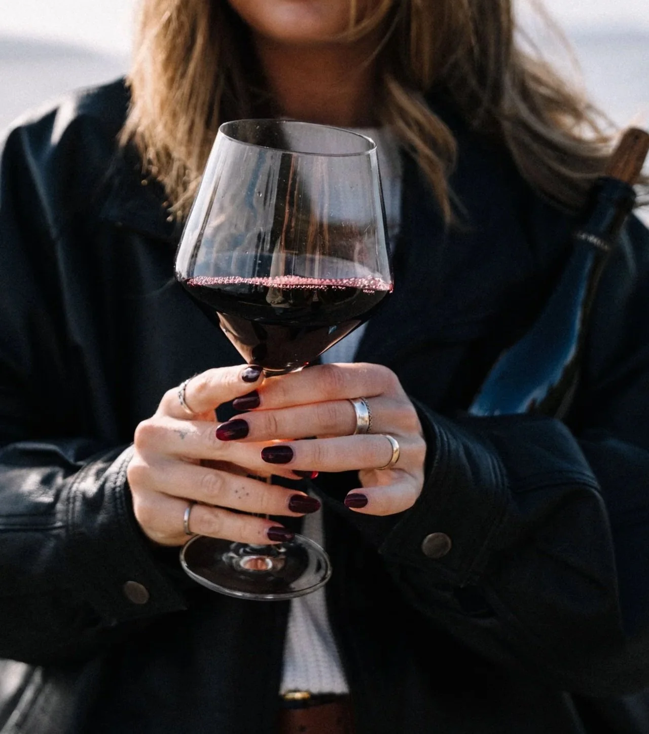 Person holding a glass of red wine with painted nails and rings, wearing a black leather jacket.
